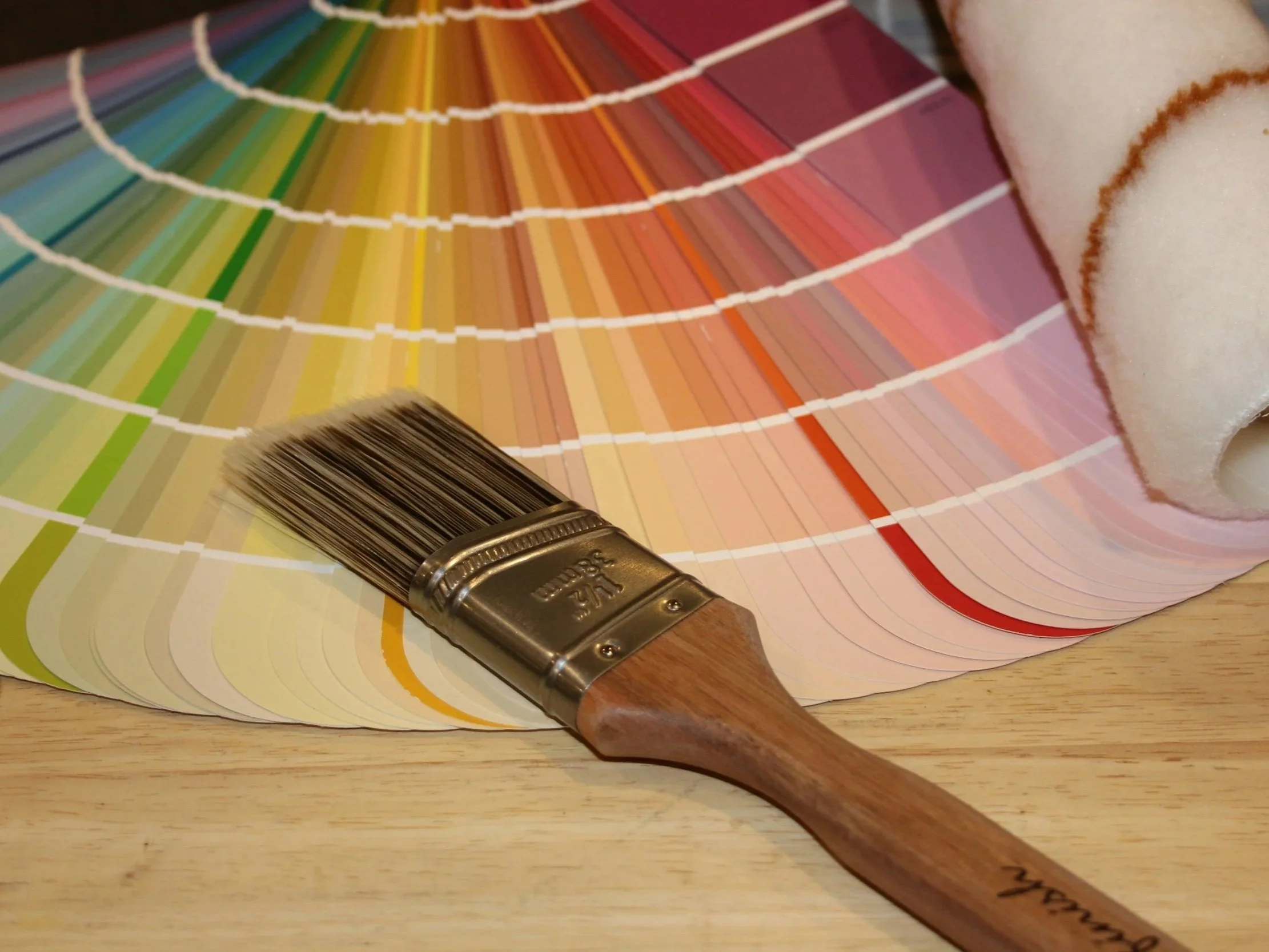 A wooden paintbrush resting on a color wheel art palette with various shades of greens, yellows, oranges, reds, and pinks, and a roll of white paper towel on a wooden surface.