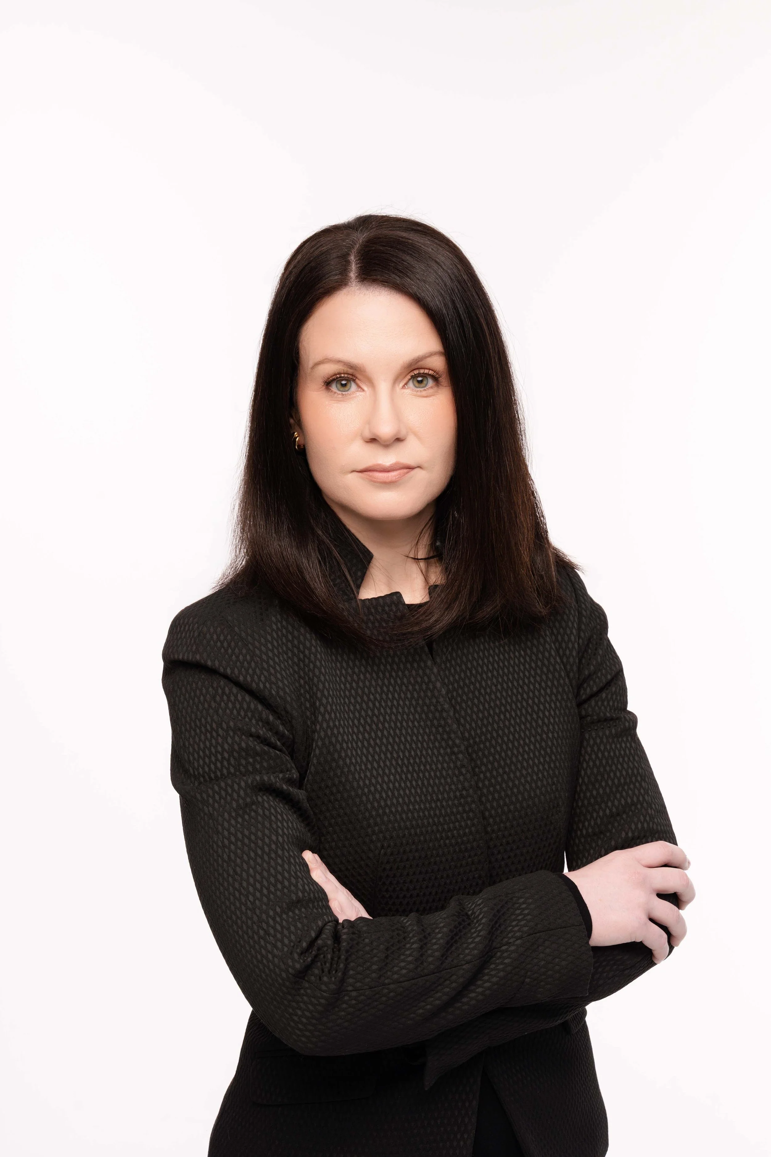 A woman with dark brown hair crossed arms wearing a black textured blazer looking at the camera with a neutral expression against a plain white background.
