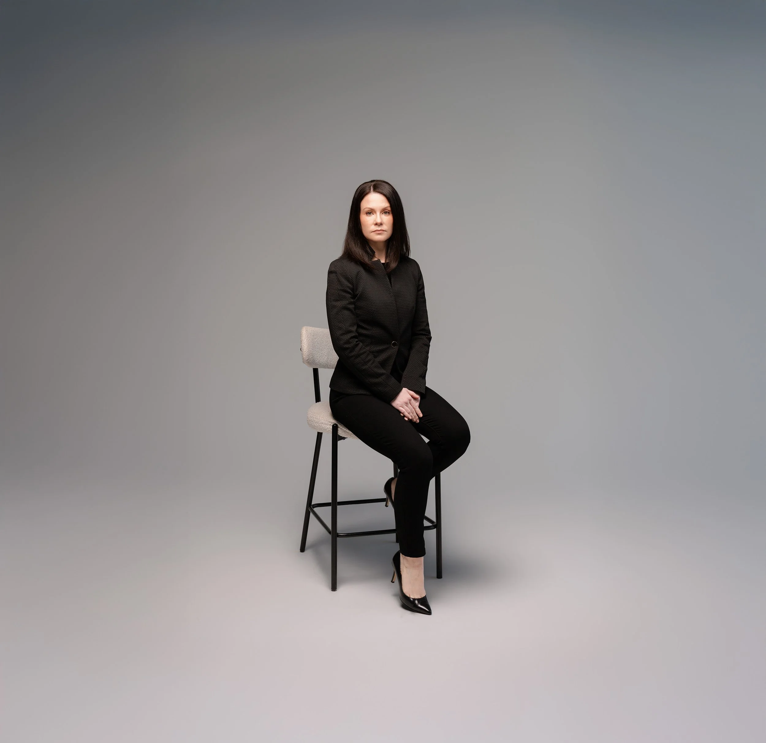 A woman with black hair sitting on a tall chair in a studio with a plain gray background, wearing a black blazer and black pants, looking directly at the camera.