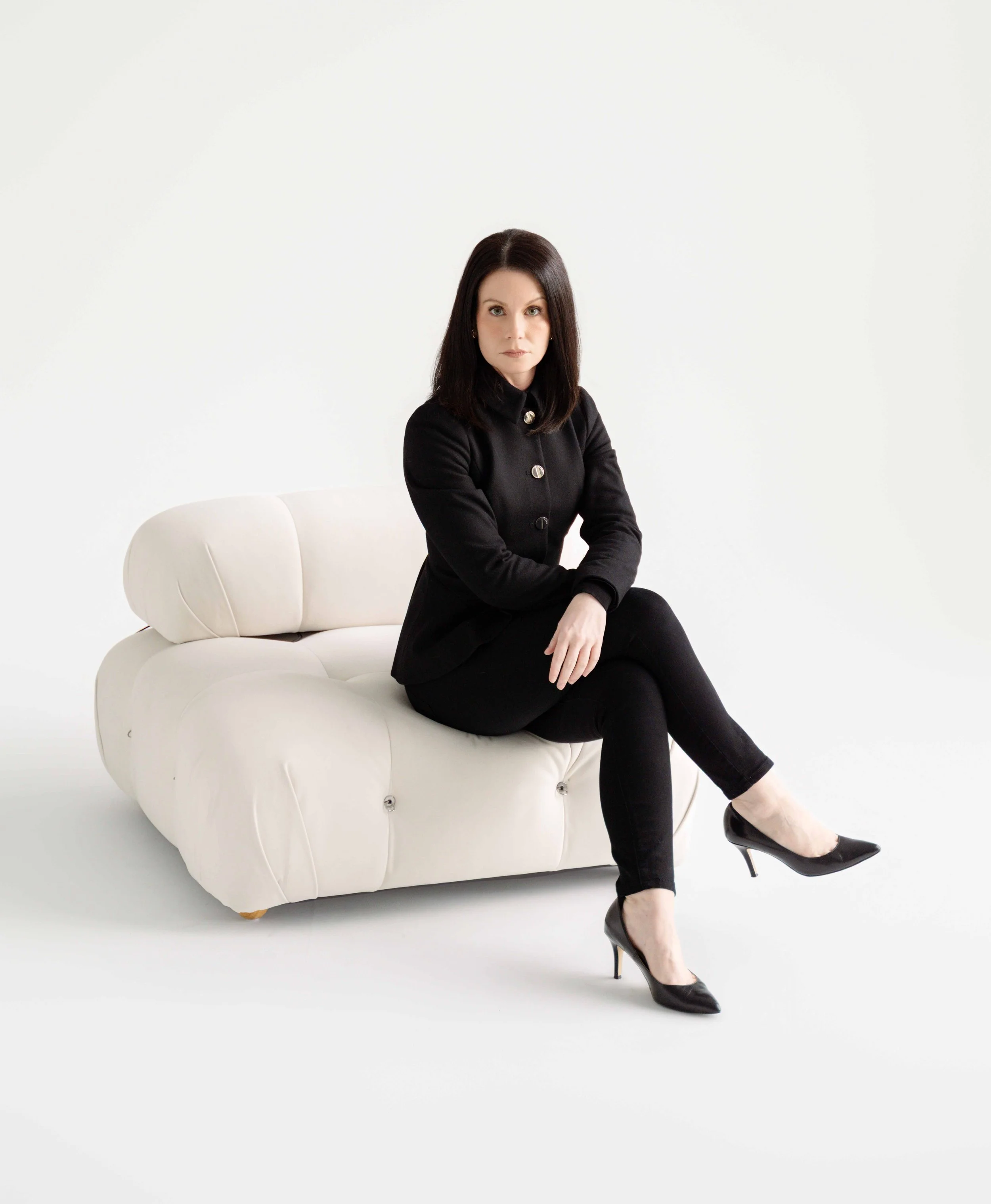 A woman wearing a black blazer and black pants sitting on a white modern sofa against a plain white background.