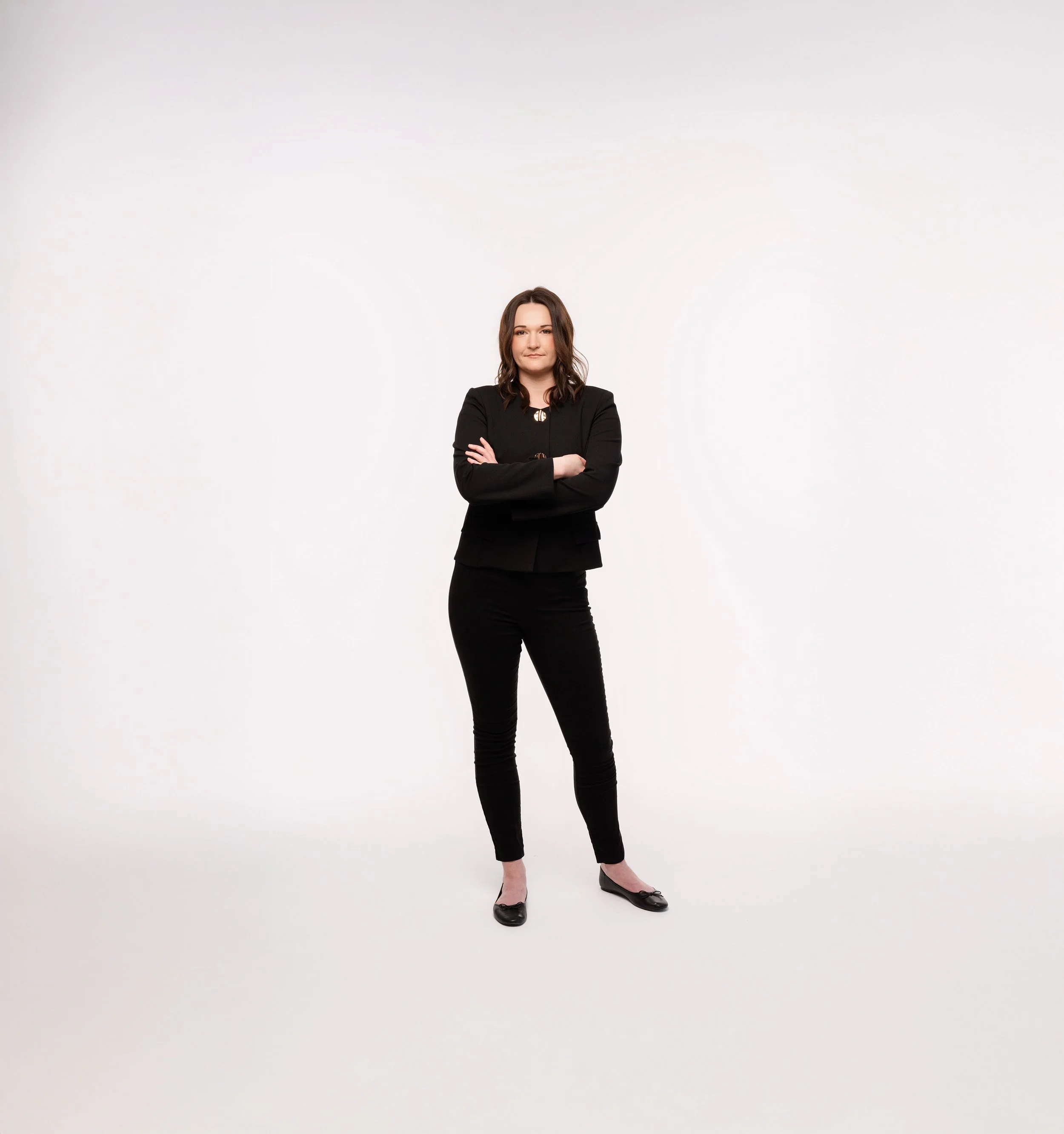 Woman with brown hair in black suit standing confidently with arms crossed, against a plain white background.