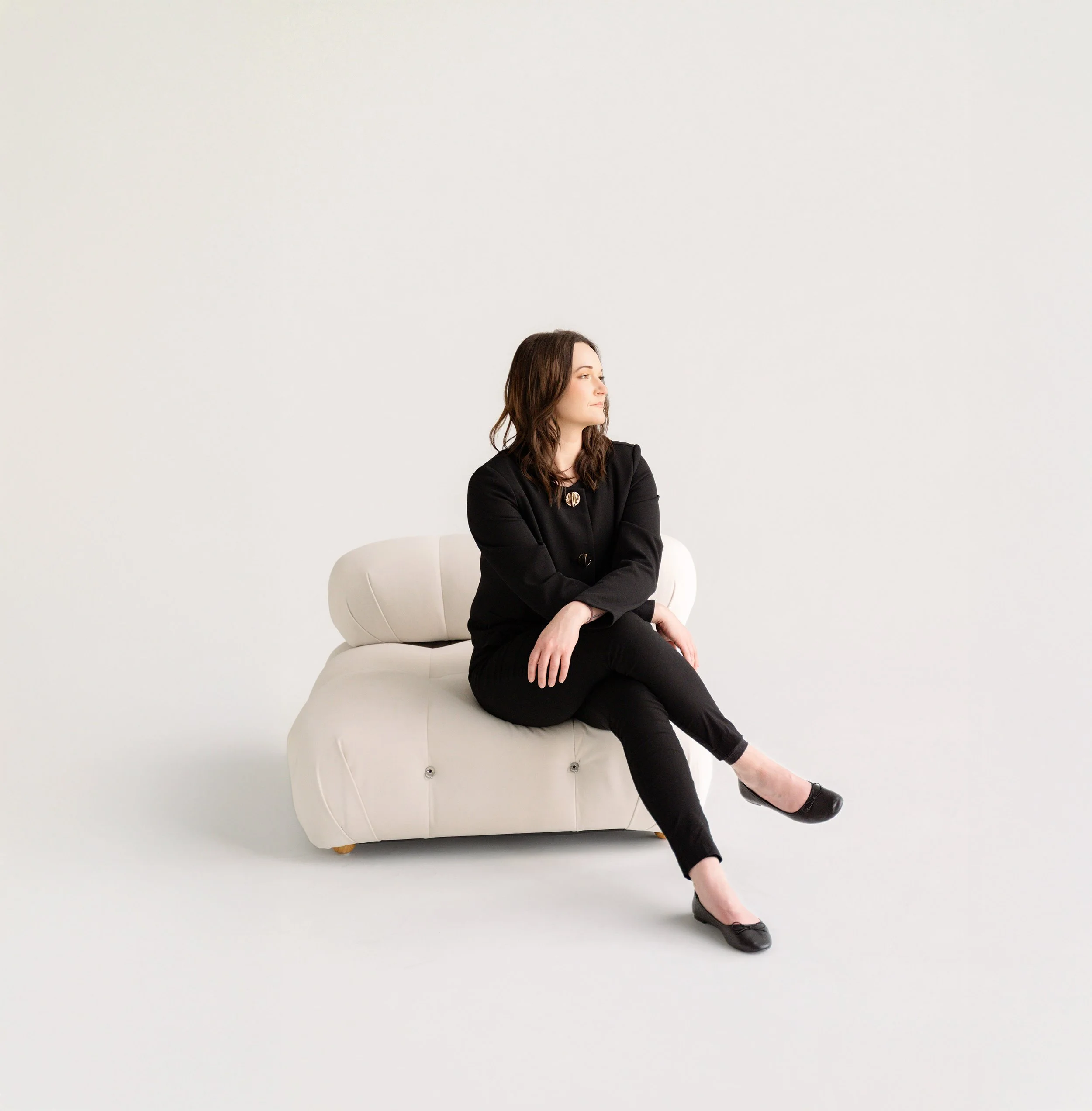 A woman with brown hair wearing a black outfit sitting on a white sofa against a plain white background.