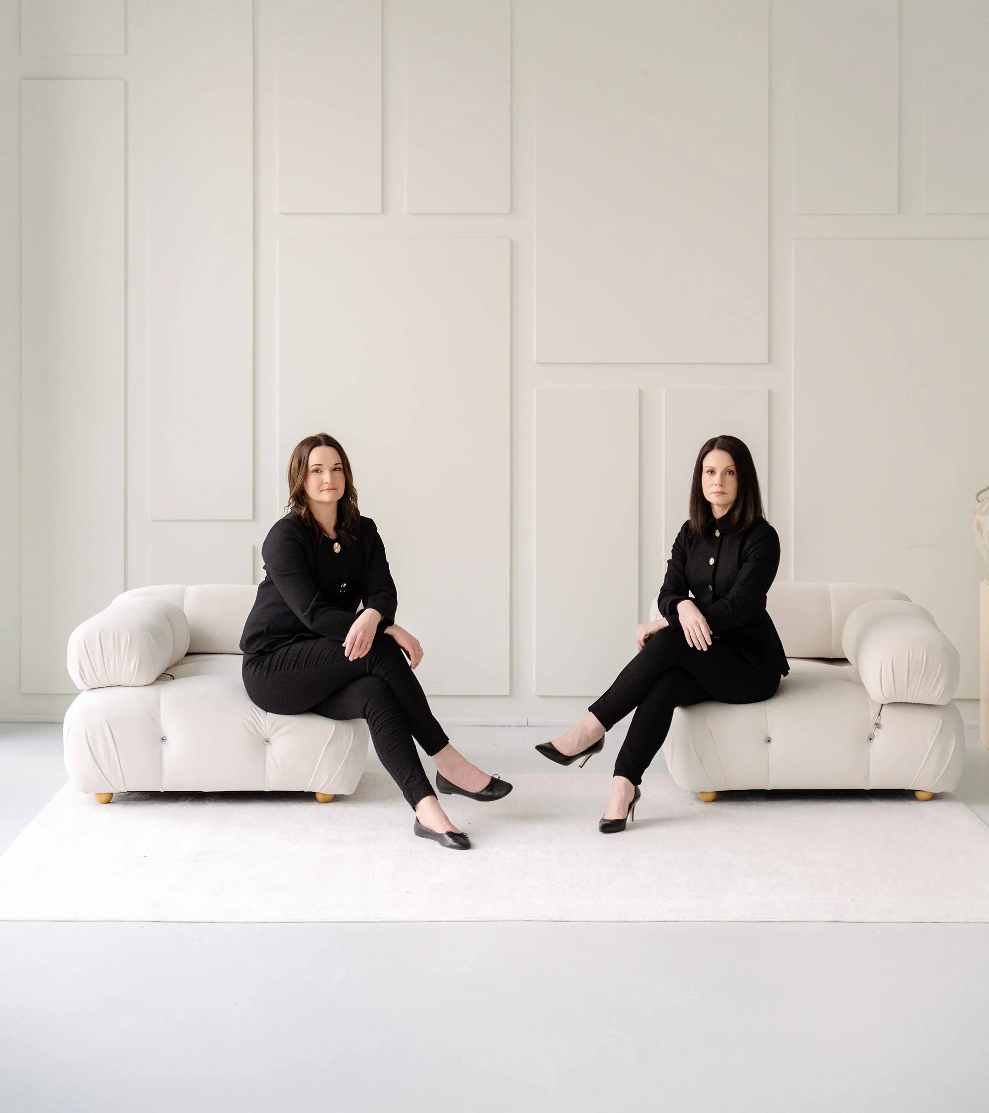 Two women seated on white couches in an all-white room, dressed in black, with a minimalist background.