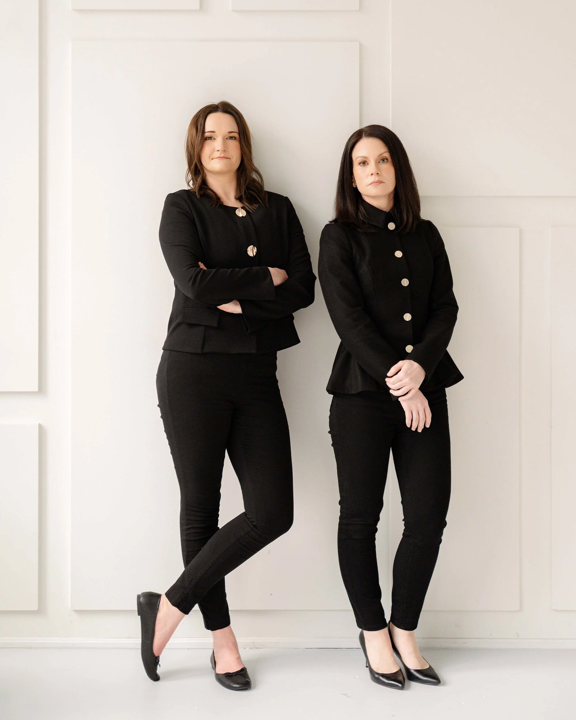 Two women standing with arms crossed, wearing black outfits, in front of a white wall with geometric panels.