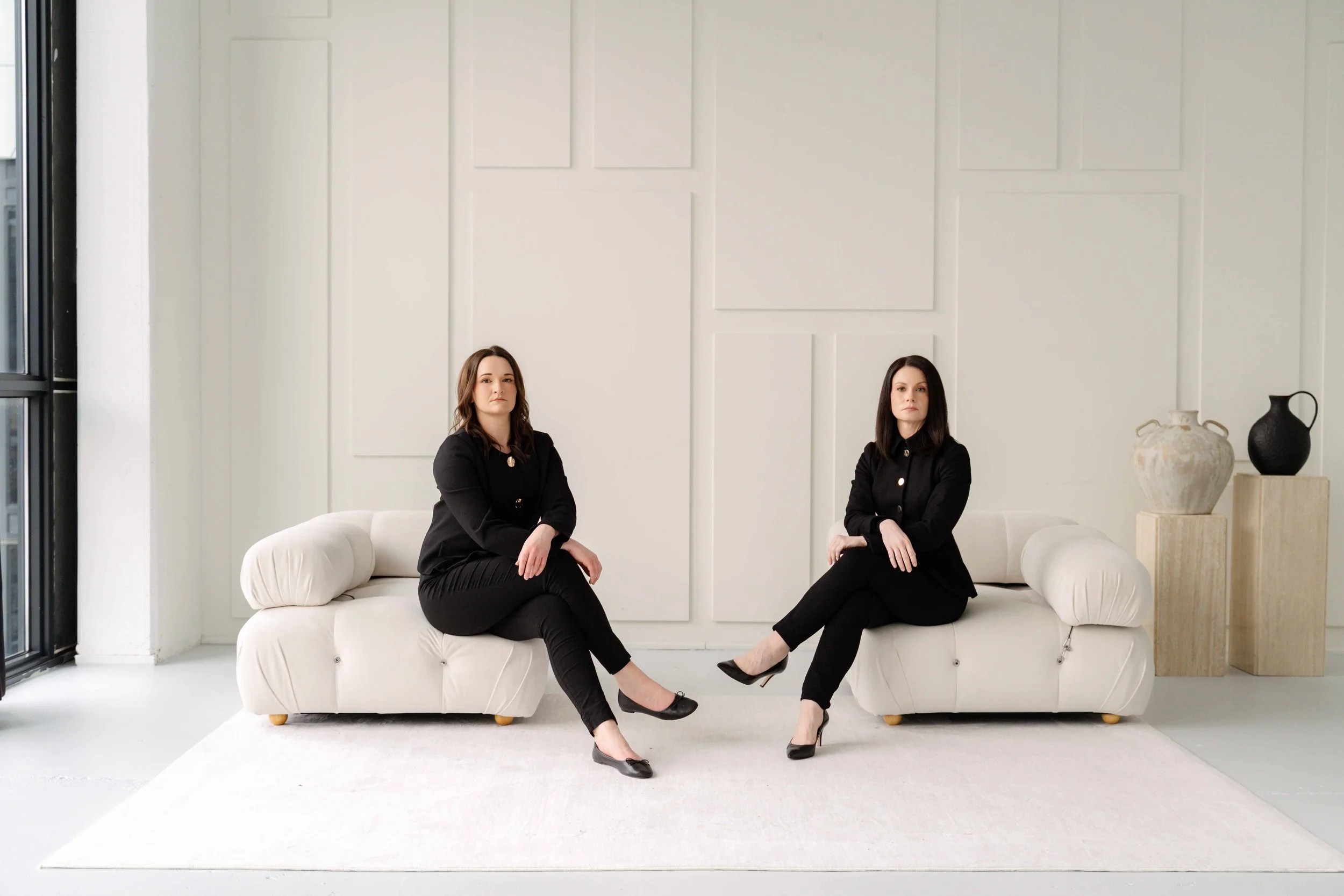 Two women with dark brown and black hair, wearing black suits and black heels, sitting on white sofas in a minimalistic room with white walls, a large window on the left, and decorative vases on the right.