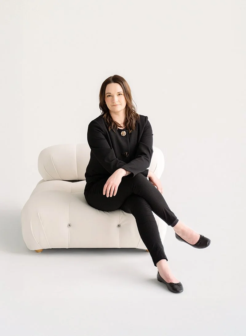 Woman sitting on a white sofa against a plain white background, wearing a black blazer and pants, with black shoes.