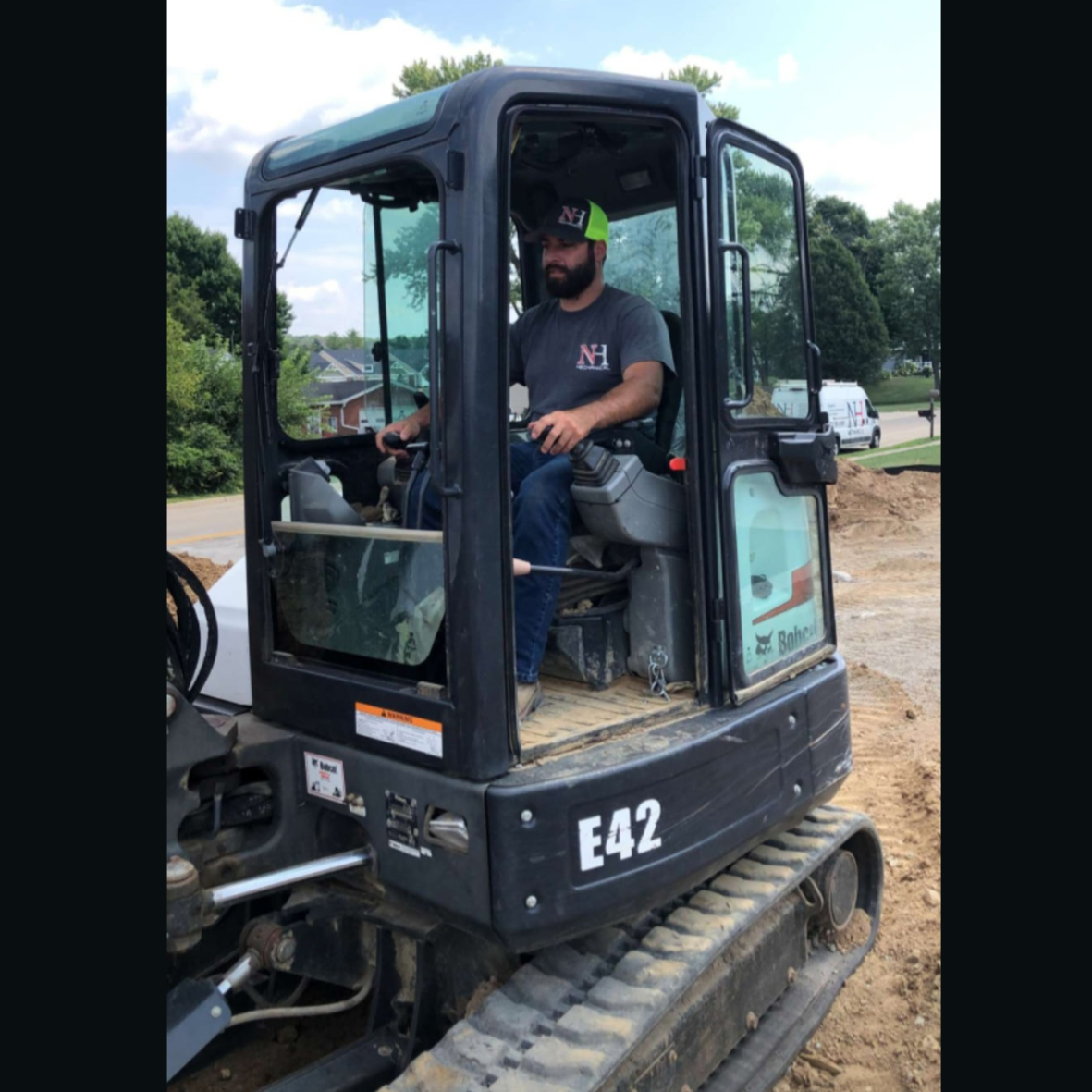 Man operating a compact excavator on a construction site, surrounded by trees and houses in the background.