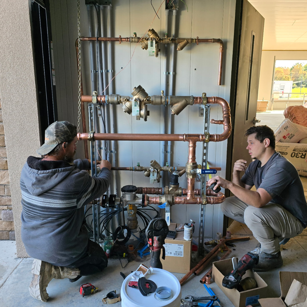 Two men working on a plumbing system with copper pipes and fittings, during construction or installation, with tools and supplies around them.