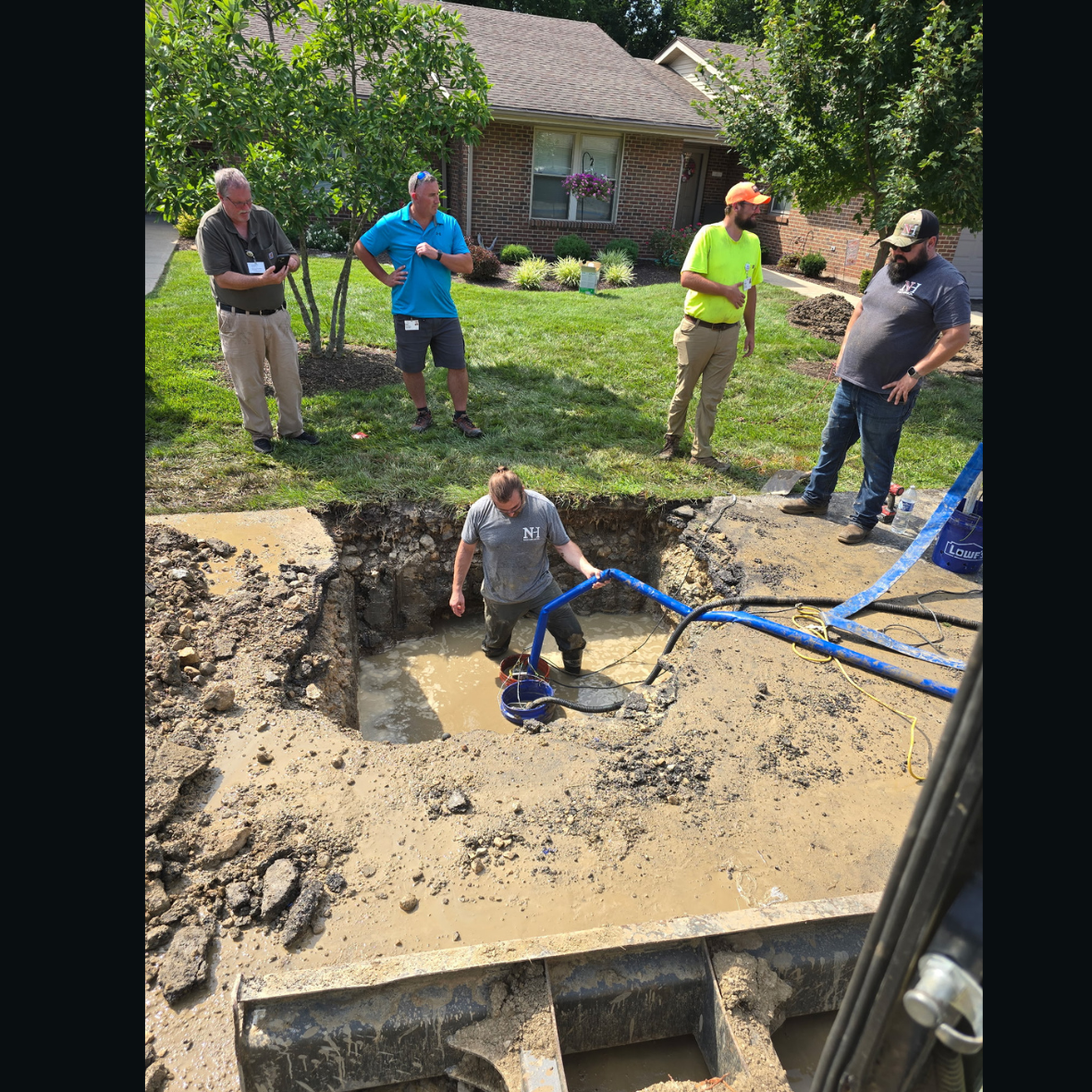 Group of men standing around a large excavation site in a residential yard, with one person inside the hole working with water and equipment. The yard has grass, trees, and a brick house in the background.