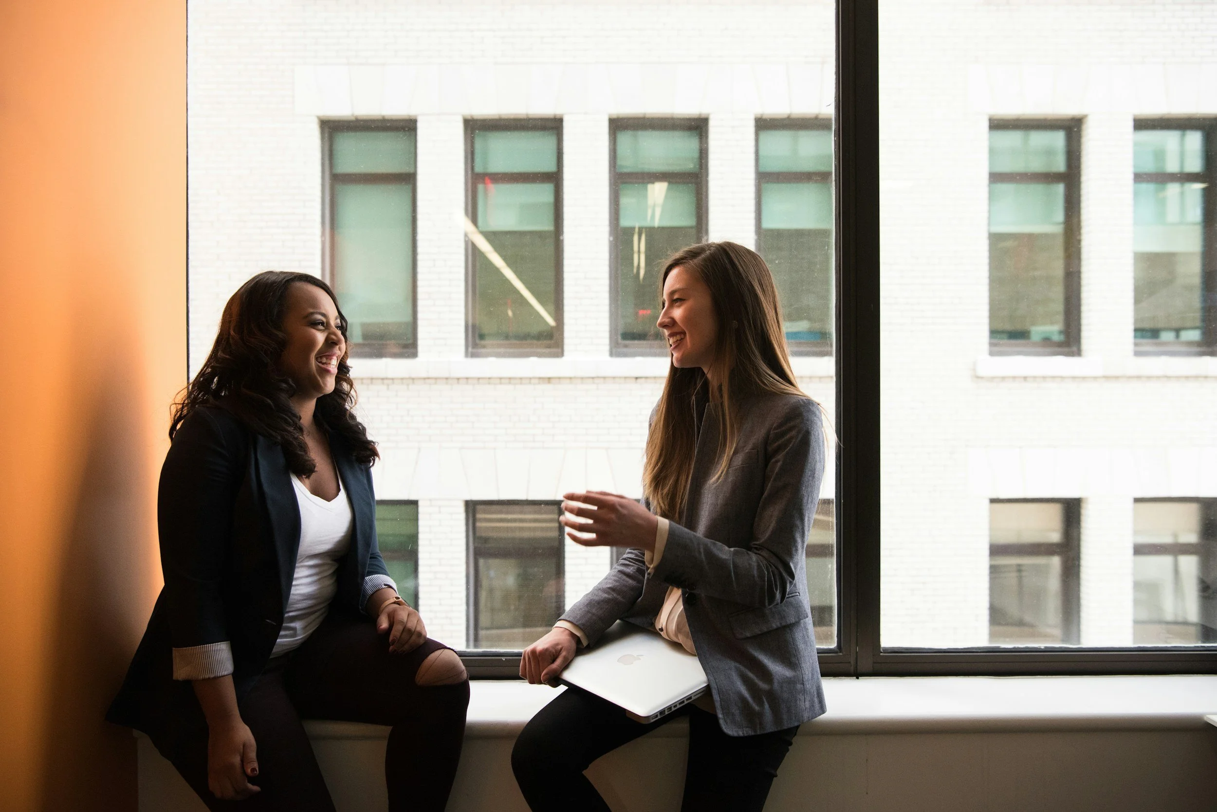 Two women are talking and smiling near a large window in a modern office building.