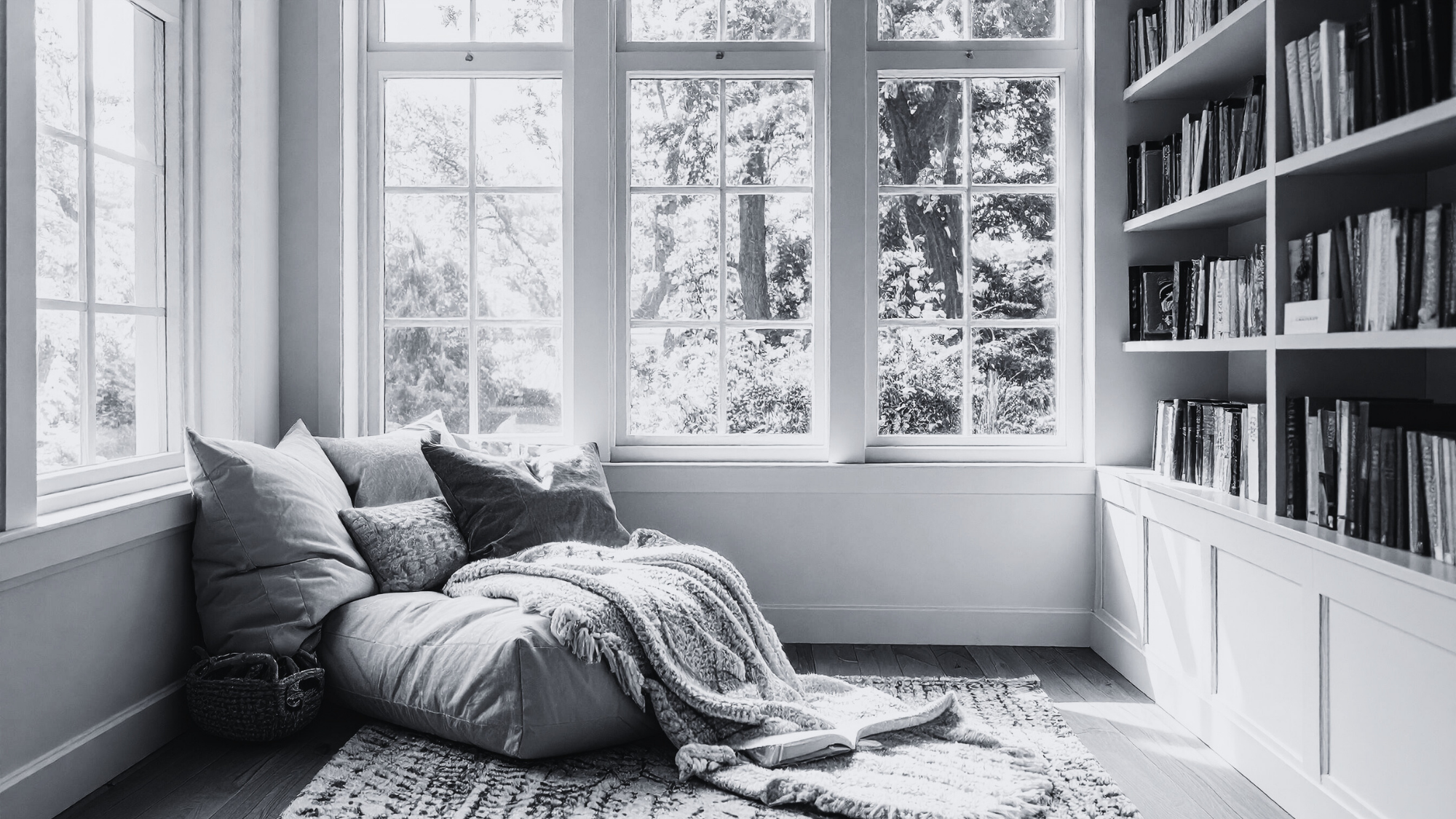 A cozy reading nook with a bean bag chair and pillows in front of large windows letting in natural light, next to a tall bookshelf filled with books.