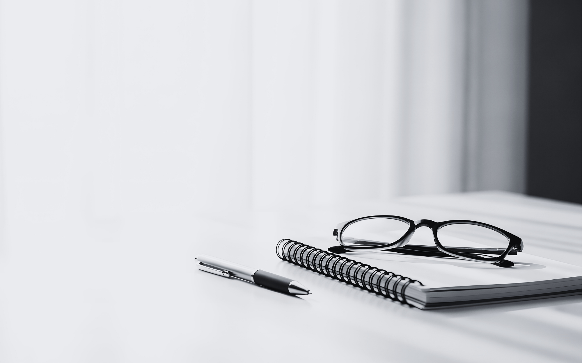 A black pen, a spiral notebook, and a pair of black-rimmed glasses on a white surface with a blurred background.