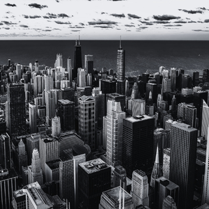 Black and white aerial view of a dense city skyline with tall skyscrapers, Lake Michigan in the background, and cloudy skies.