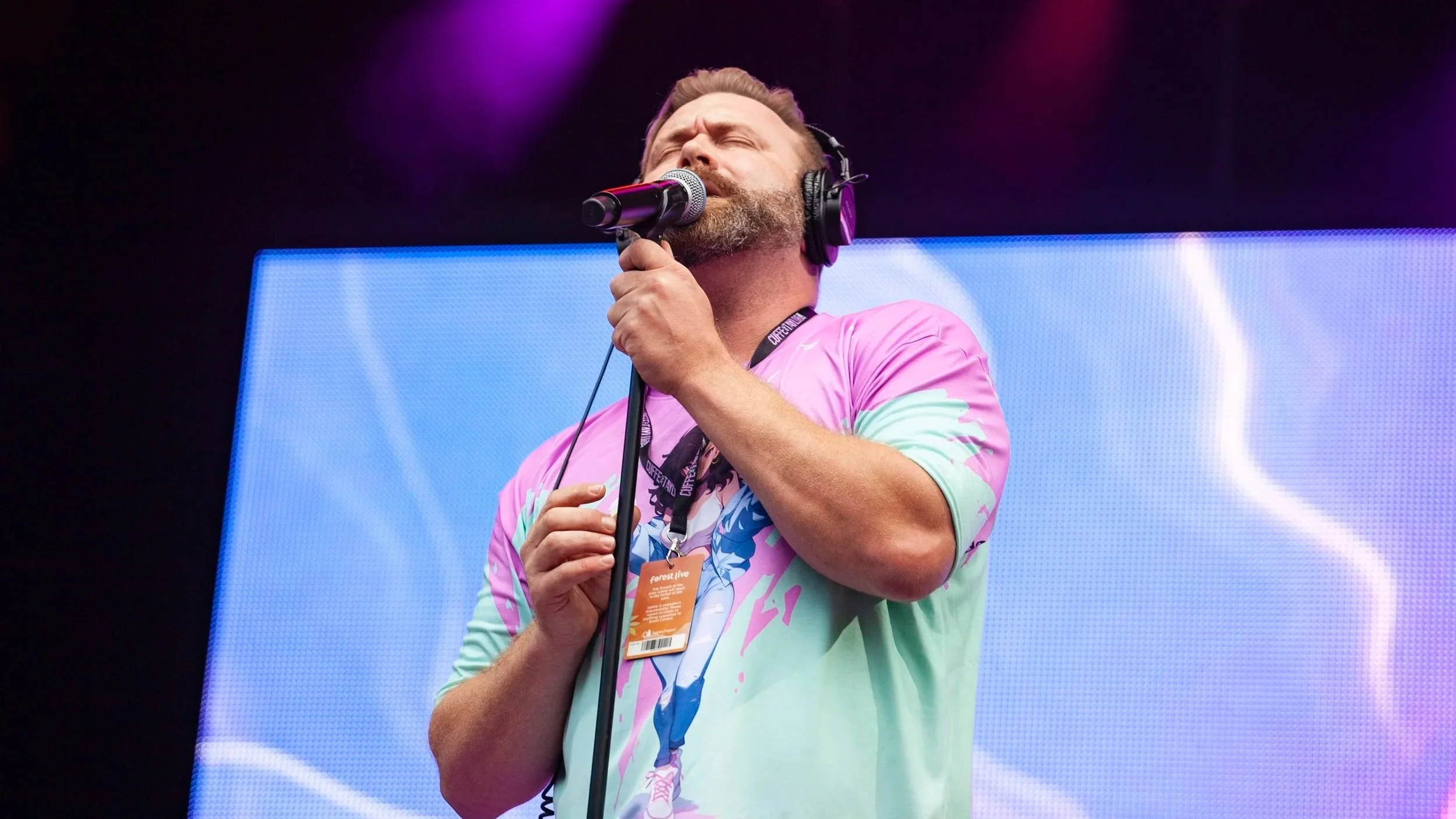 Daniel Beddingfield, with a beard, singing into a microphone, wearing headphones, and a colourful graphic T-shirt, standing in front of a large digital screen with blue and purple visuals.