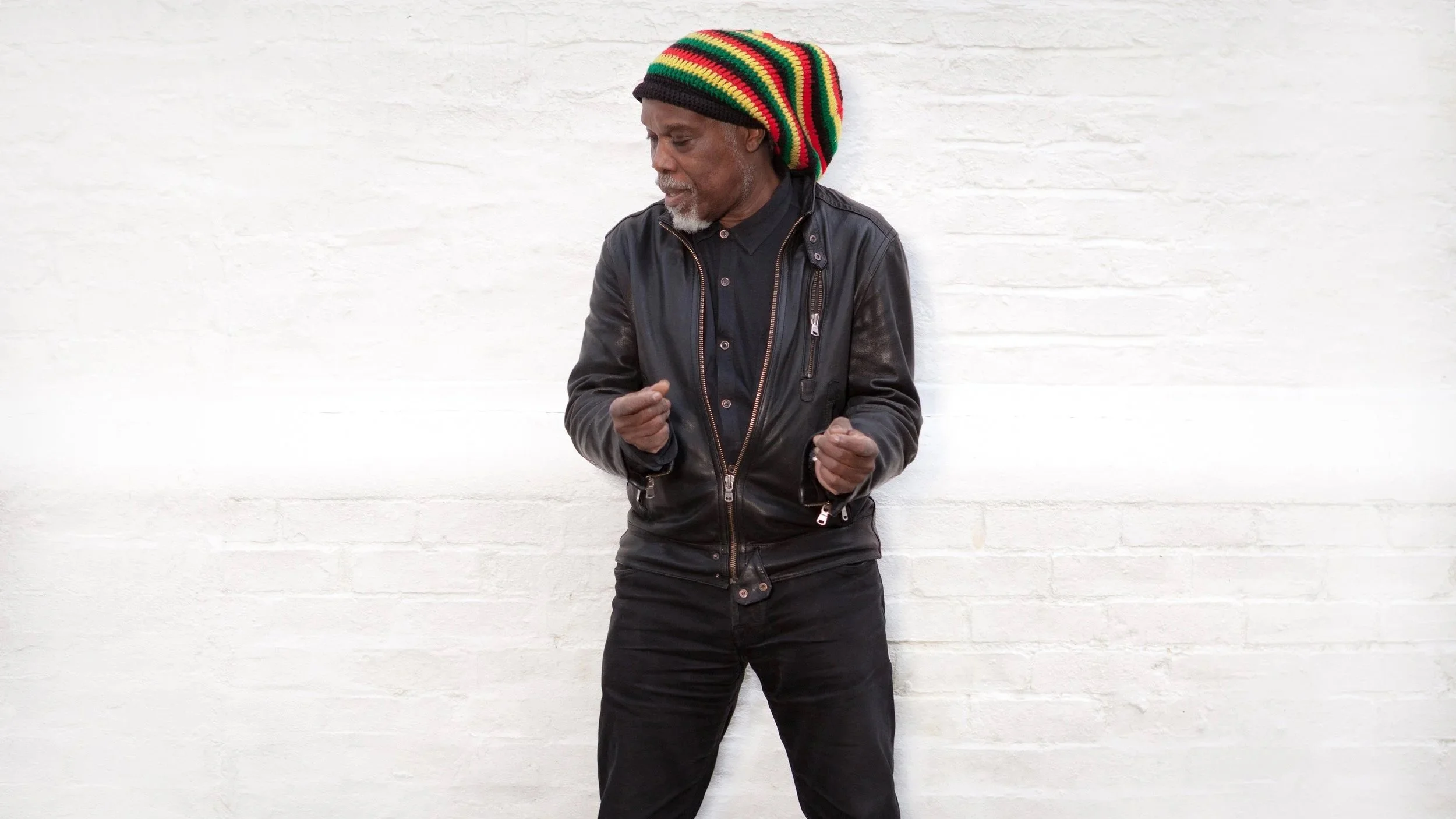 Billy Ocean, wearing a black leather jacket, black shirt, and black pants, standing against a white brick wall, with a red, green, and yellow striped knit cap, looking down, fists clenched.