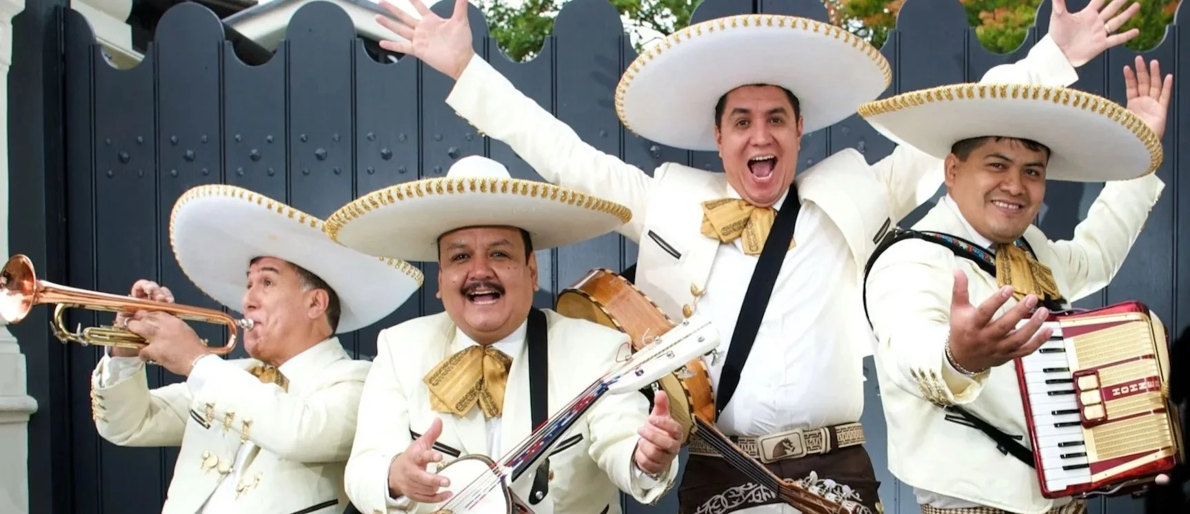 Group of musicians wearing traditional Mexican mariachi costumes with large sombreros, playing instruments and smiling outside in front of a dark fence.