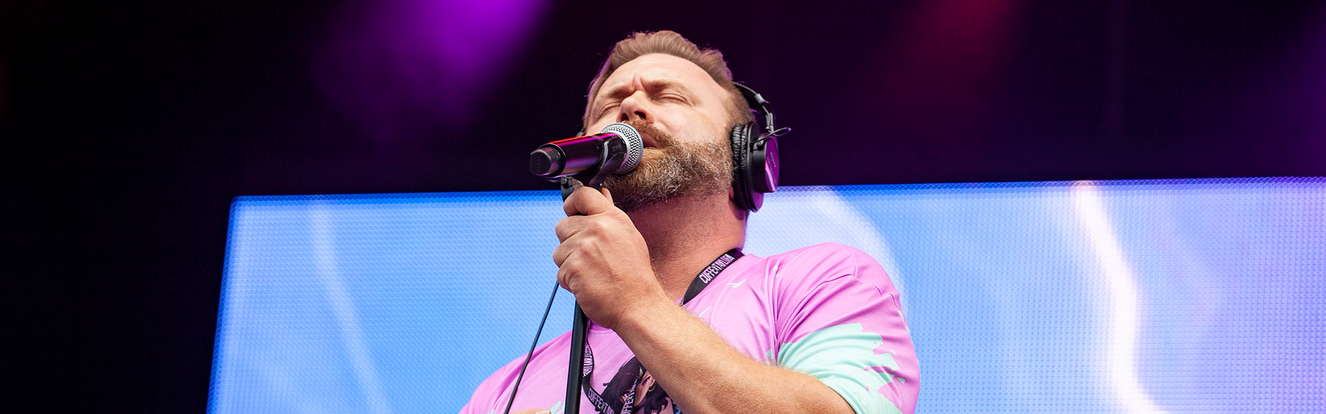 A man with a beard singing into a microphone on stage with purple and blue lighting, wearing a pink shirt and headphones.
