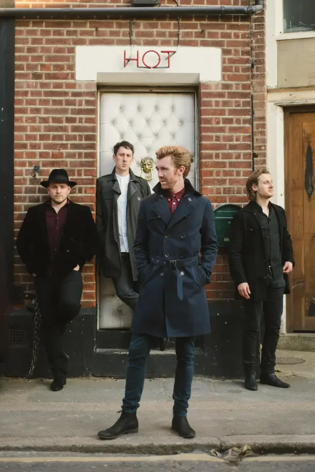 Four members of the band, The Milk, dressed in dark clothing, standing against a brick wall, with a sign above that reads 'HOT' and a small decorative skull behind them.