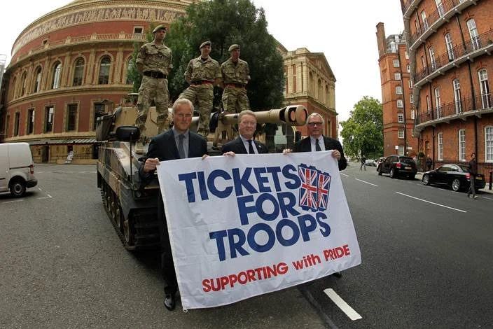 Three men stand in front of a tank on a city street, holding a large sign that reads 'Tickets for Troops, Supporting with Pride.' Behind them, three soldiers in camouflage military uniforms sit atop the tank, with buildings and parked cars in the bac