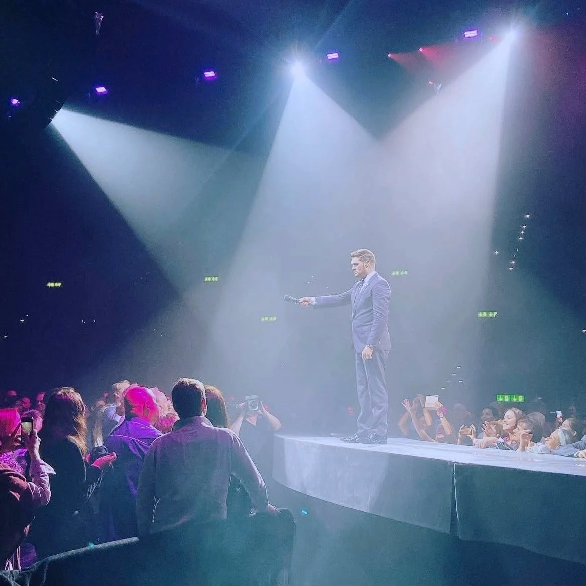 A man in a suit on a stage with bright spotlights, holding a microphone, with an audience watching and taking photos.
