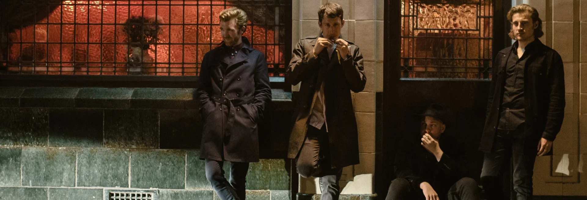 Four men in dark clothing standing and sitting against a brick wall with barred windows at night.