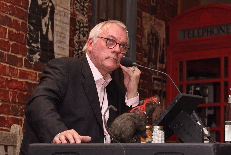 A man with glasses and white hair, dressed in a black suit, sits at a table with a microphone, looking thoughtful. On the table, there's a small model or figurine, a computer monitor, and various items. Behind him is a brick wall with a black-and-whi