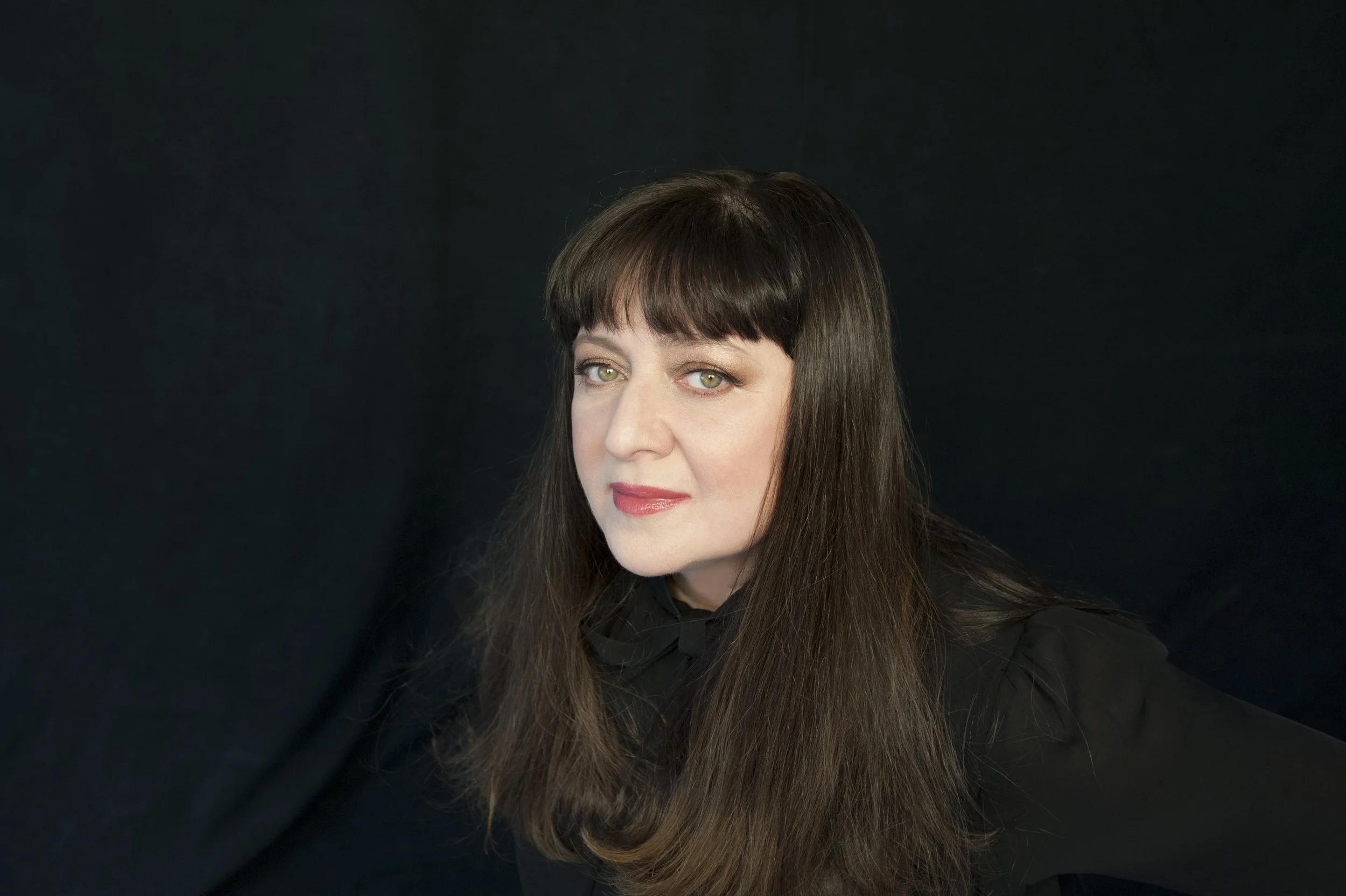 A woman with long brown hair and green eyes, wearing a black top, looks directly at the camera against a black background.