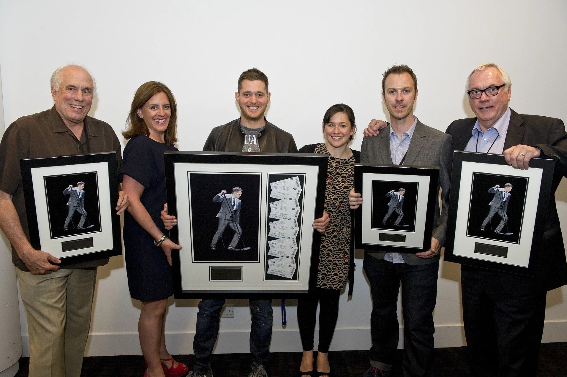 Group of seven people holding framed photos and awards, posing for a picture against a plain wall.
