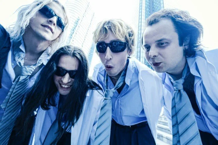 The four members of Bilderbuch, wearing sunglasses and white shirts, look down at the camera with serious faces, tall buildings in the background, and a clear sky.