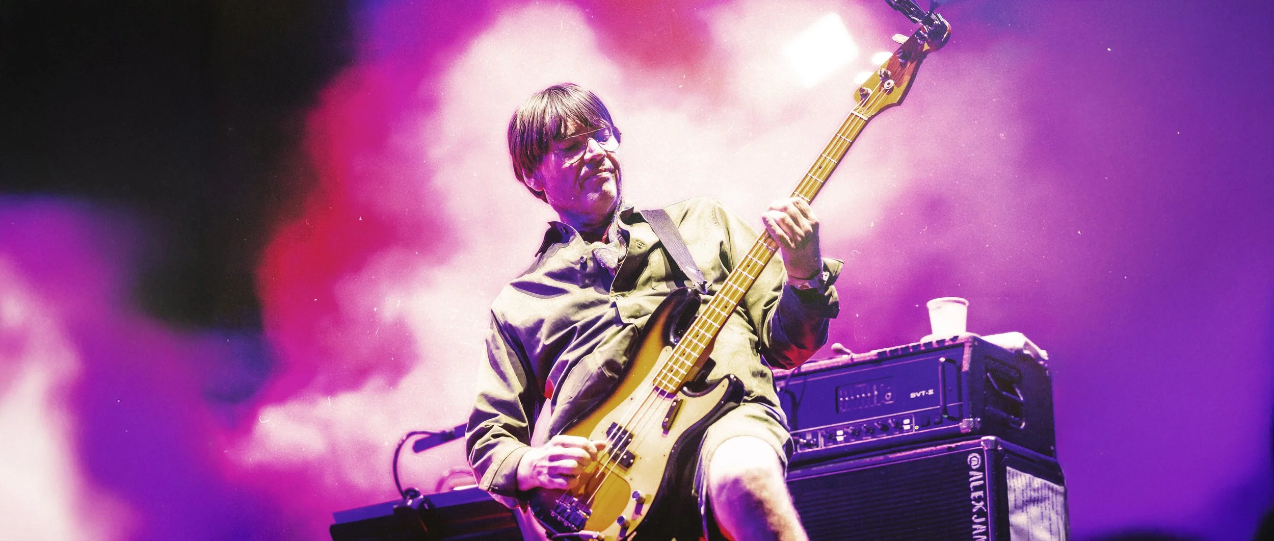 Alex James', playing a bass guitar on stage with purple and pink lighting, wearing glasses and a khaki shirt, with an amplifier and a cup on top of it nearby.