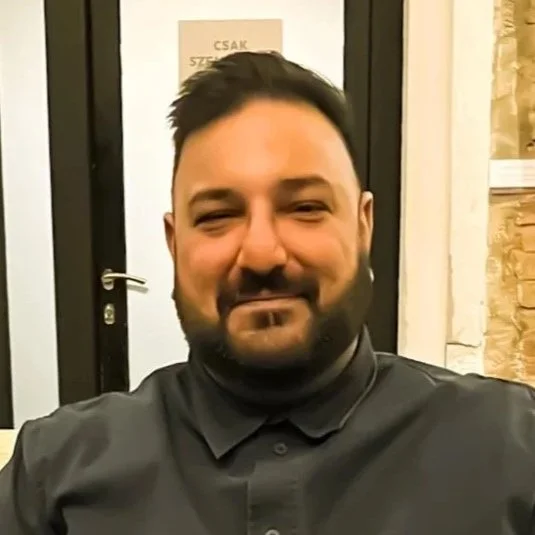 South Asian Male, sitting on a white sofar chair, with a microphone in his hand as he is speaking. Wering a black tshirt, with cream trousters and white & blue shoes.