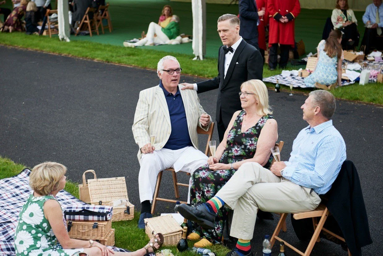People at an outdoor gathering, some sitting and some standing on a paved surface with picnic blankets and baskets around. One man in a striped blazer and glasses is speaking while others listen, smiling.