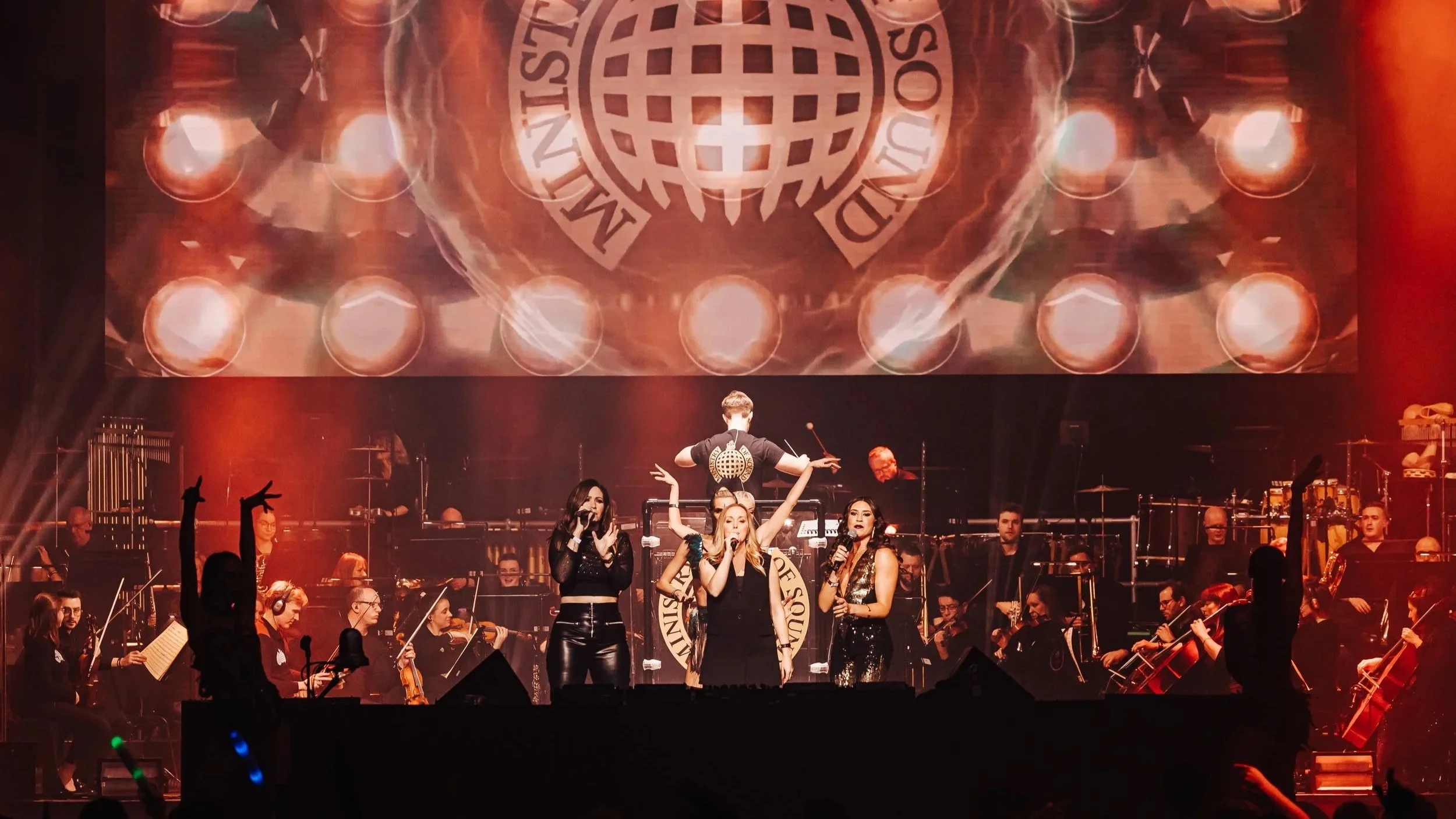 Stage scene with three female singers performing, an orchestra with string and percussion instruments behind them, and a conductor in the background. A large screen behind displays a logo with a globe and the words 'International Sound'. The stage is lit with orange and red lighting.