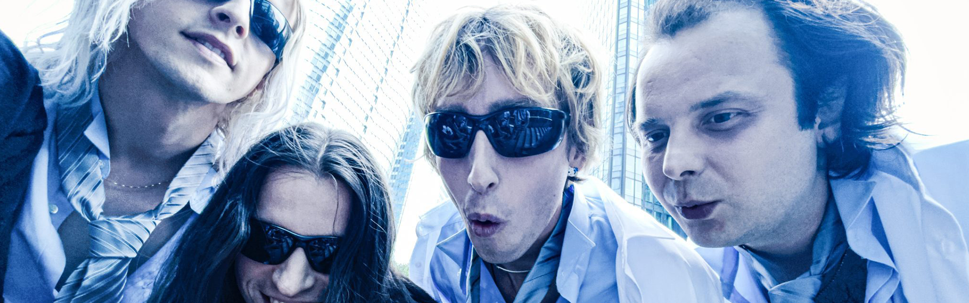 A group of five young people looking down at the camera, with modern glass buildings in the background, appearing as if they are peering through a window or camera lens.