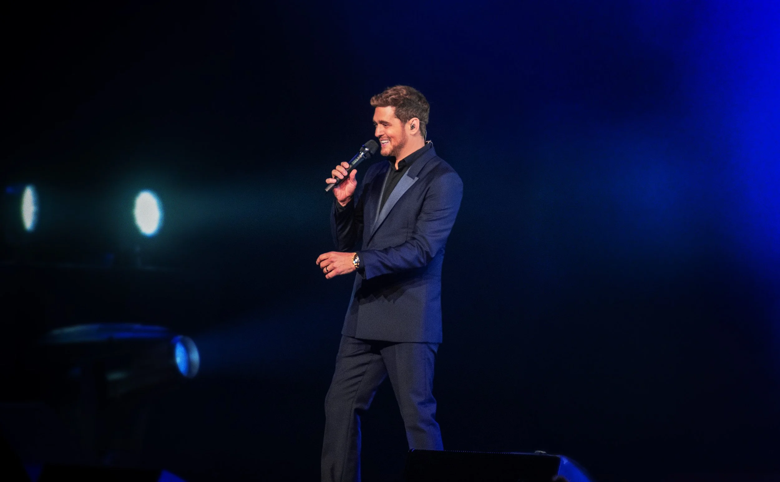 Michael Bublé in a dark blue suit holding a microphone on stage, smiling with a dark background and blue spotlight.