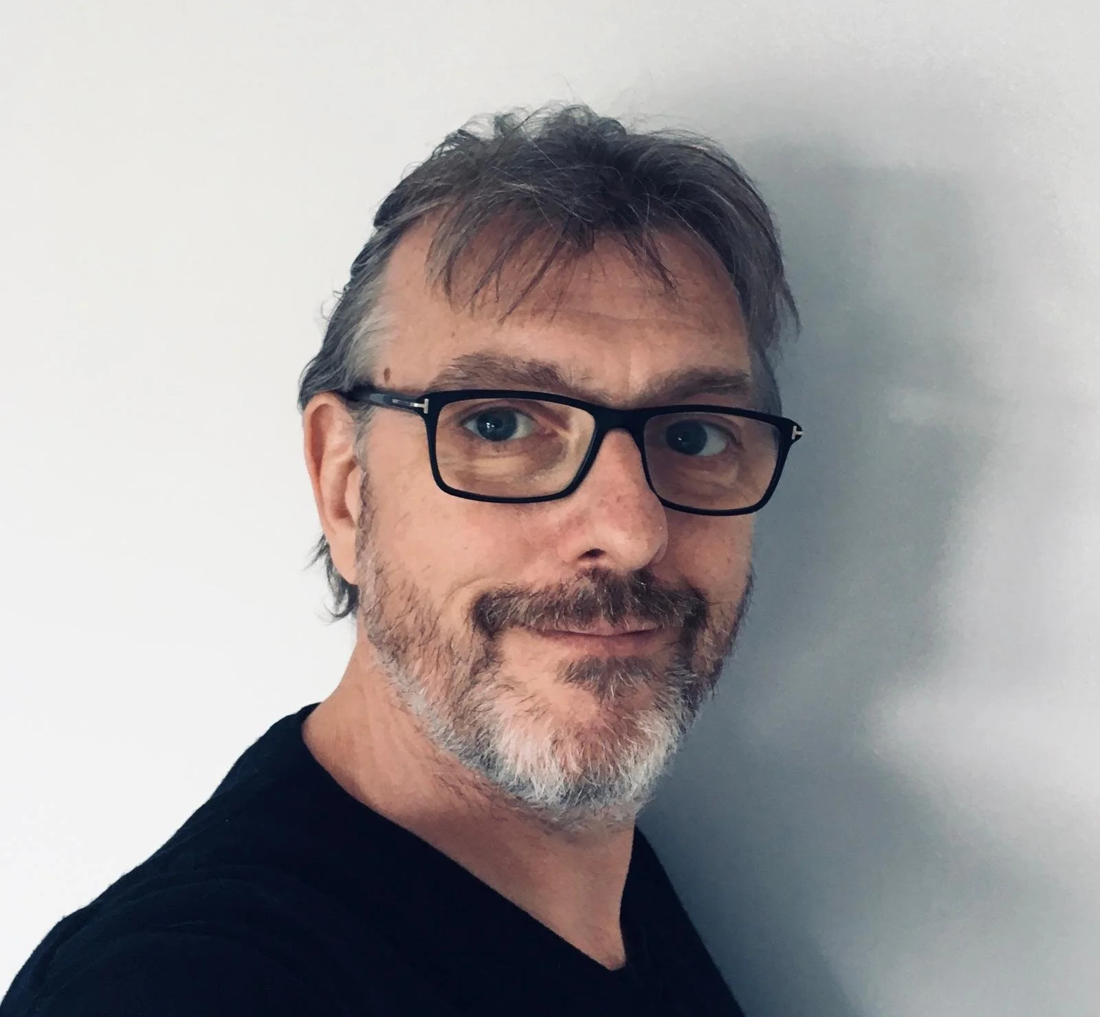 A man with curly dark hair, glasses, and a beard, wearing a dark sweater and a white shirt, sitting indoors with a plant in the background.