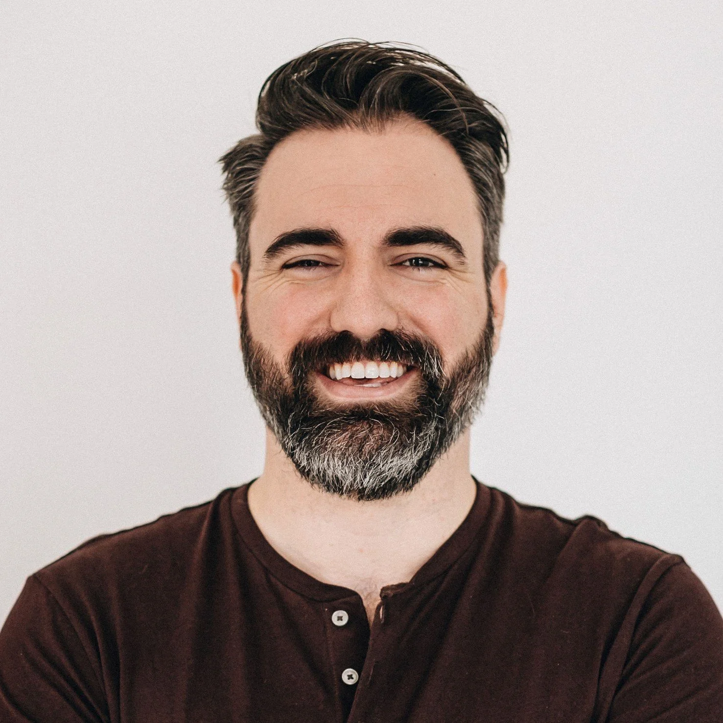 A smiling man with a beard and dark hair, wearing a dark maroon shirt, standing against a plain light-colored background.