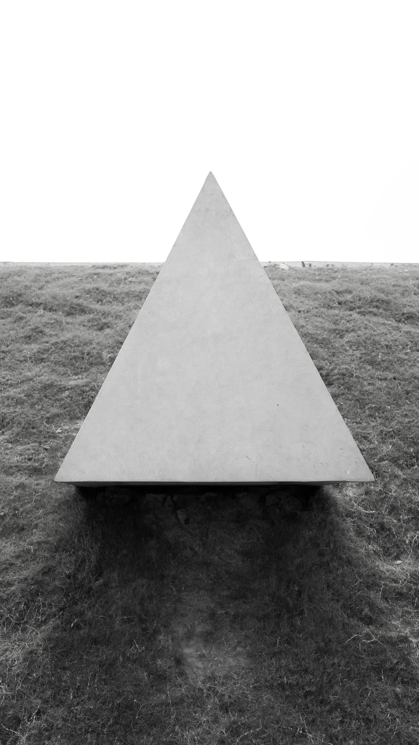 A large triangular concrete sculpture placed on grass in an outdoor setting. The background is bright, suggesting overcast weather.
