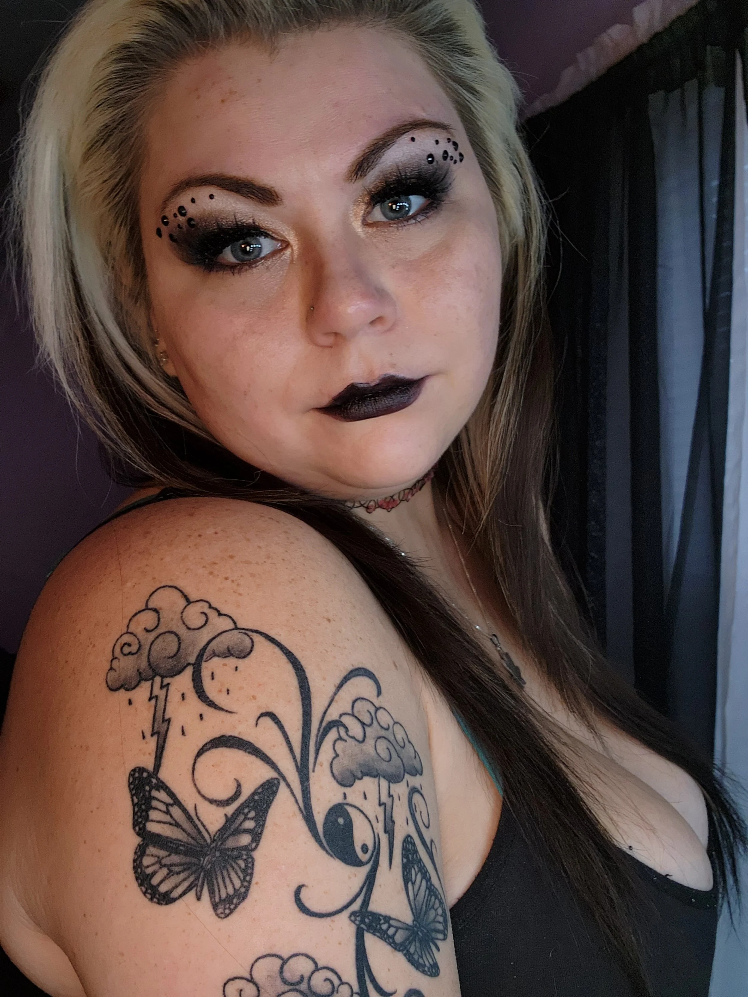 A woman with platinum blonde and dark brown hair, wearing dramatic eye makeup with black dots, dark lipstick, and a tattoo of butterflies and clouds on her shoulder.