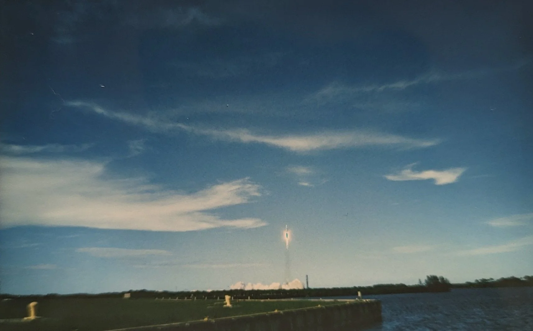 A rocket launch with a rocket ascending into the sky, leaving a bright trail of smoke and fire, against a backdrop of blue sky with wispy clouds and a body of water in the foreground.