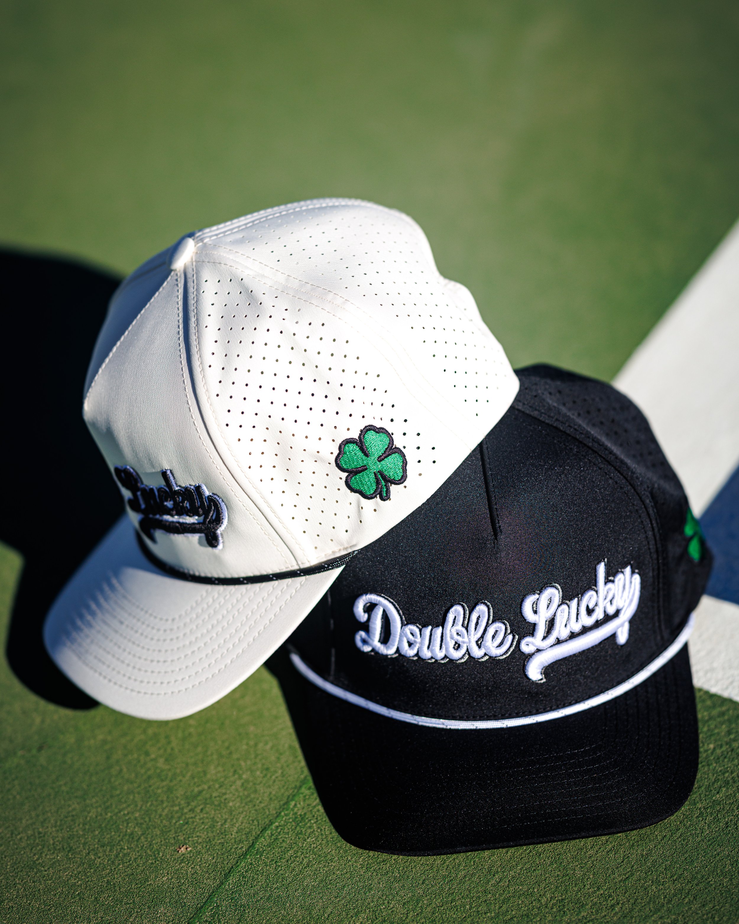 Two baseball caps, one white with a green shamrock and the word 'Lucky' in black, the other black with white cursive lettering saying 'Double Lucky' and a small green shamrock, placed on a green surface.