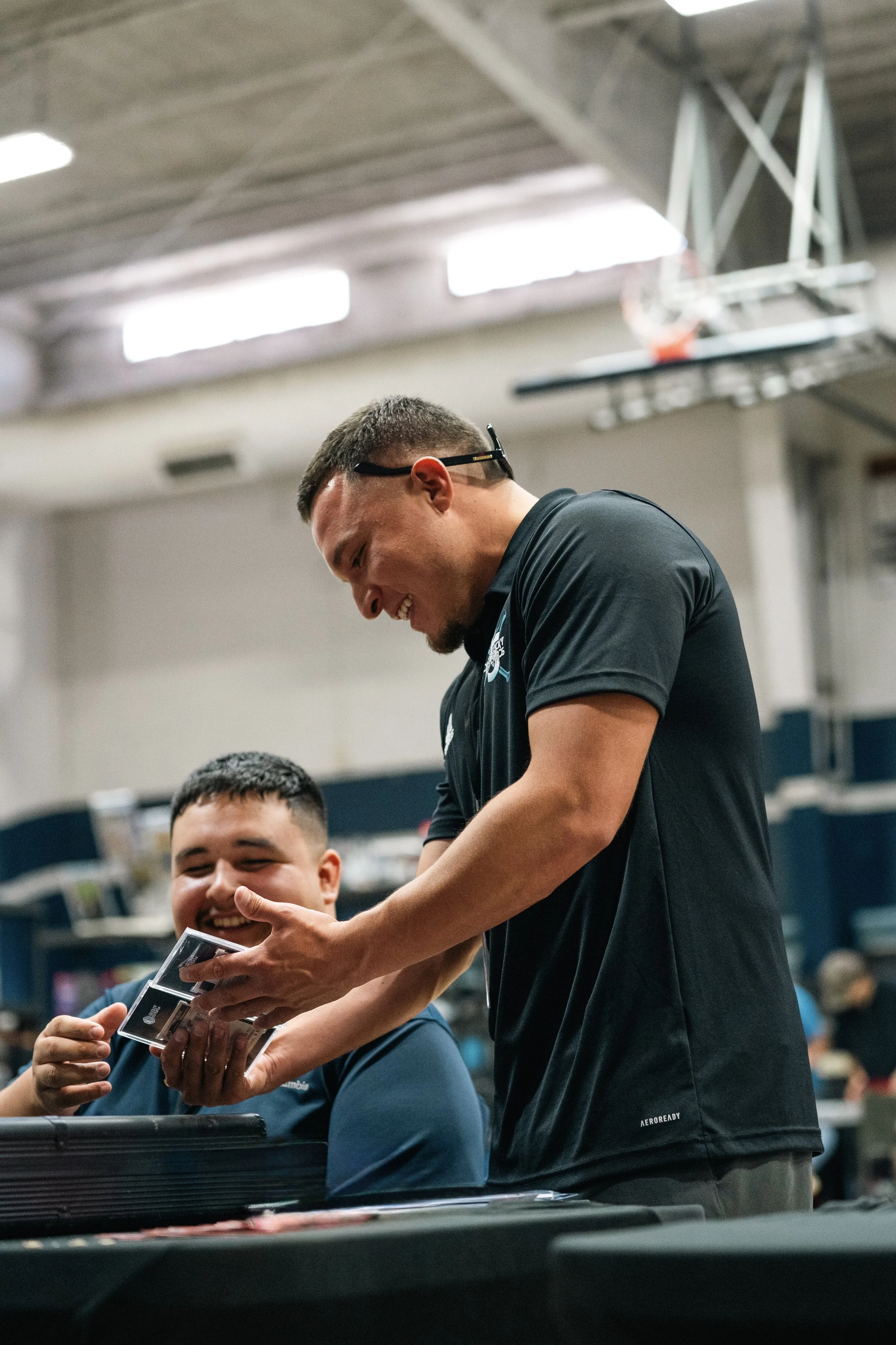 Two young men in a gym, one smiling and sitting at a table, the other standing and handing him a small box, possibly a gift or a prize.