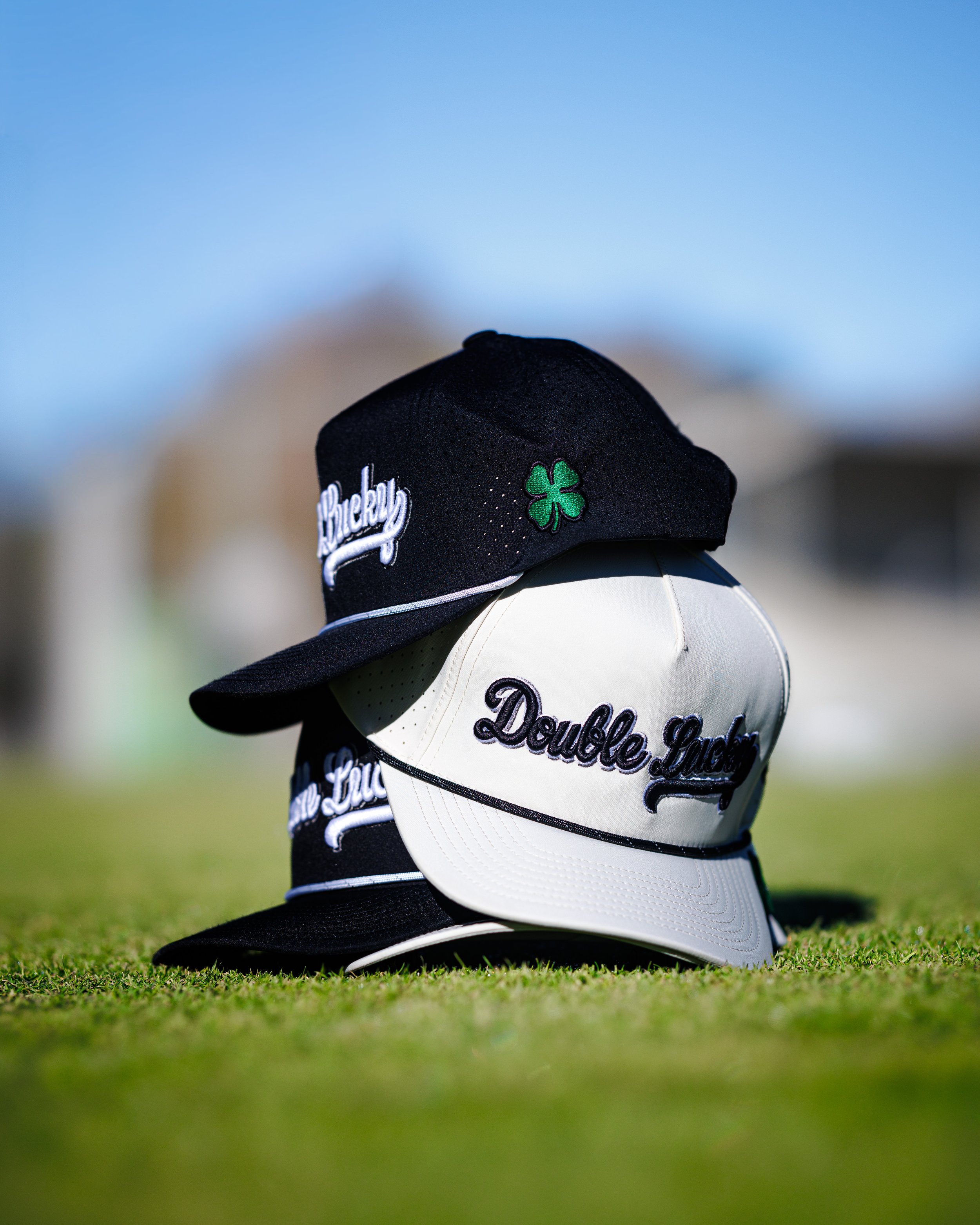 Three golf caps, two black and one white, placed on green grass with a blurred outdoor background. The caps have embroidery, including a four-leaf clover and the words 'Lucky' and 'Double Lucky'.