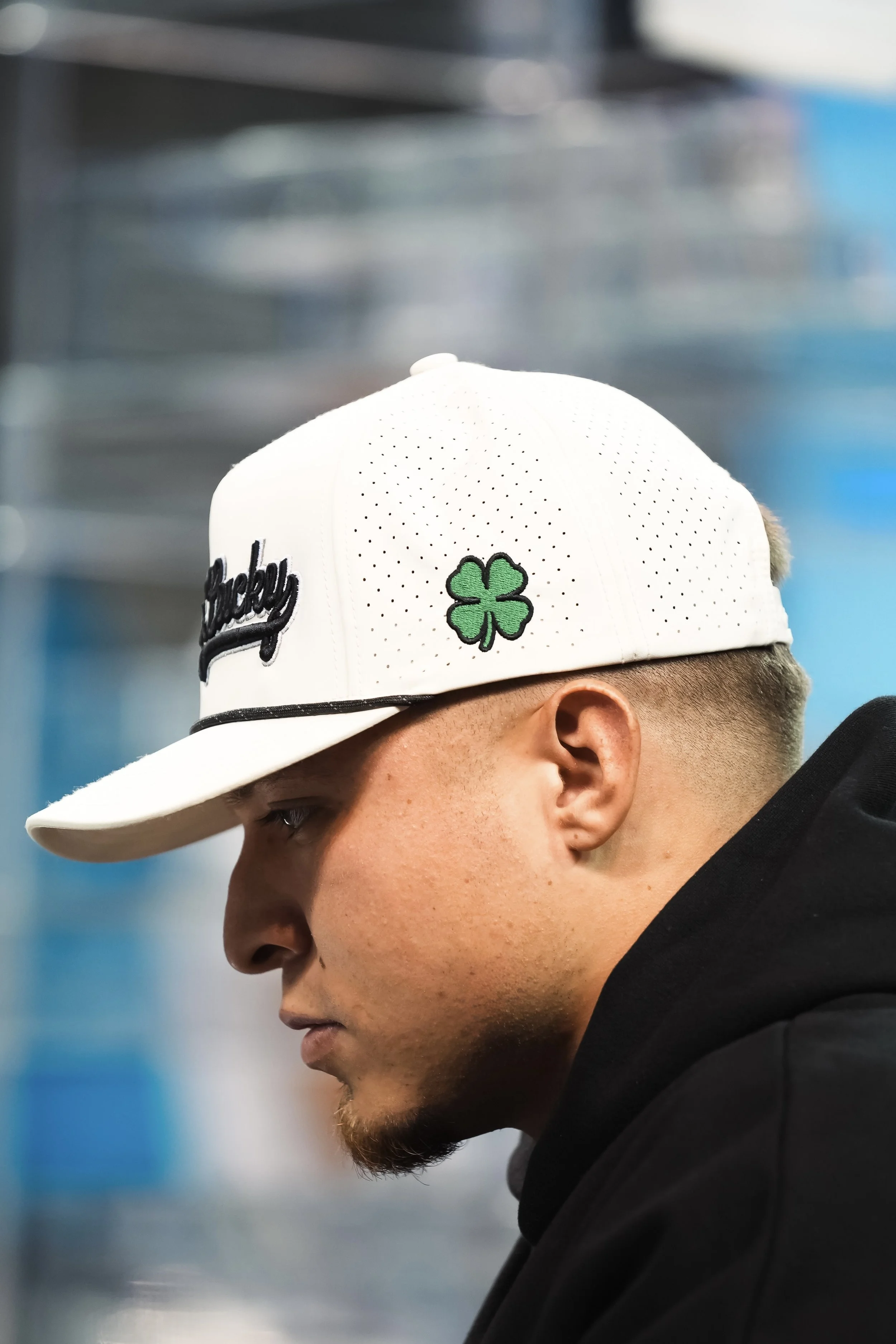 Close-up of a young man wearing a white baseball cap with a green four-leaf clover and the word 'Lucky' embroidered on it, with a blurred industrial or warehouse background.
