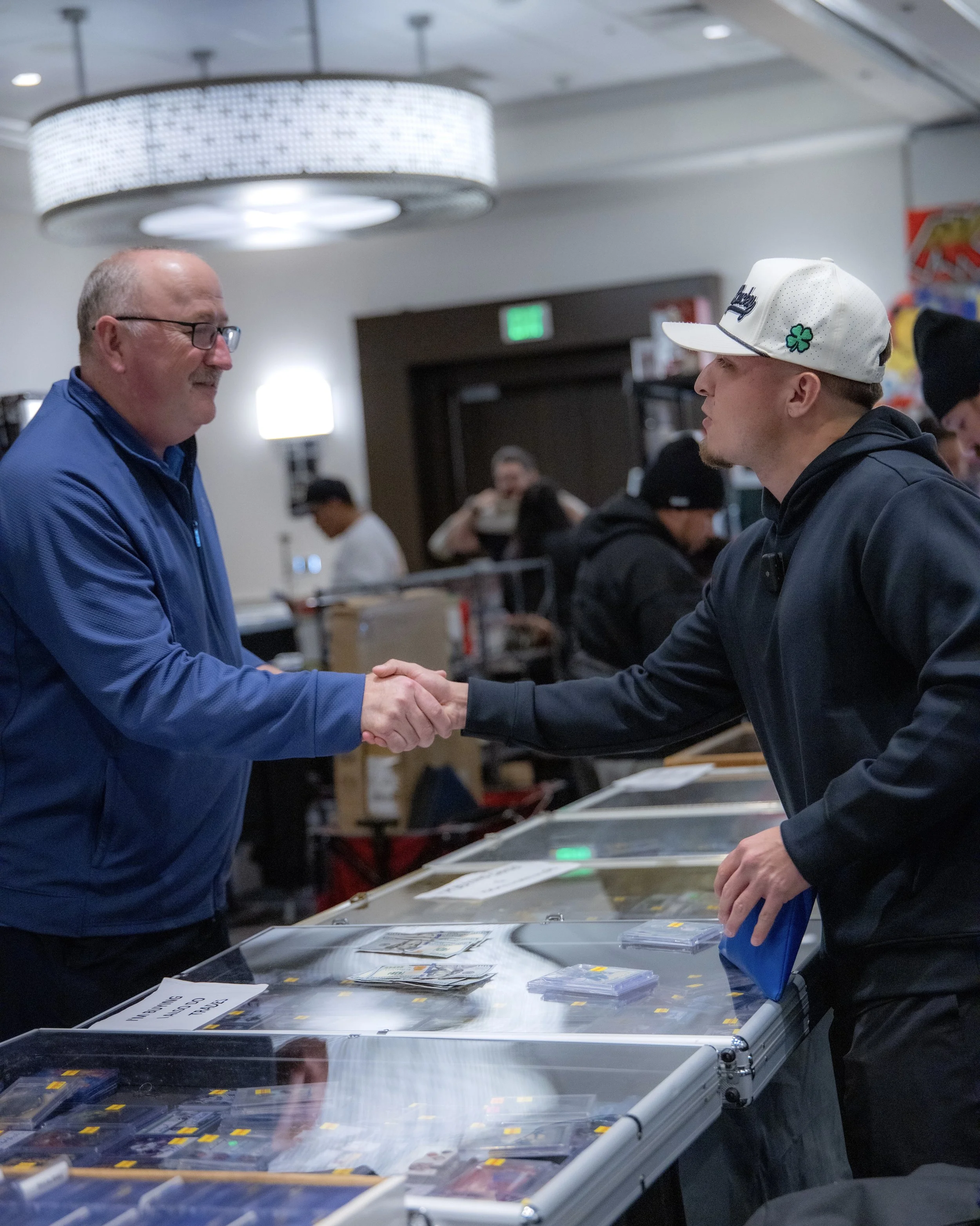 Two men shaking hands at a card trading event behind a table with trading cards, in a room with other people in the background.