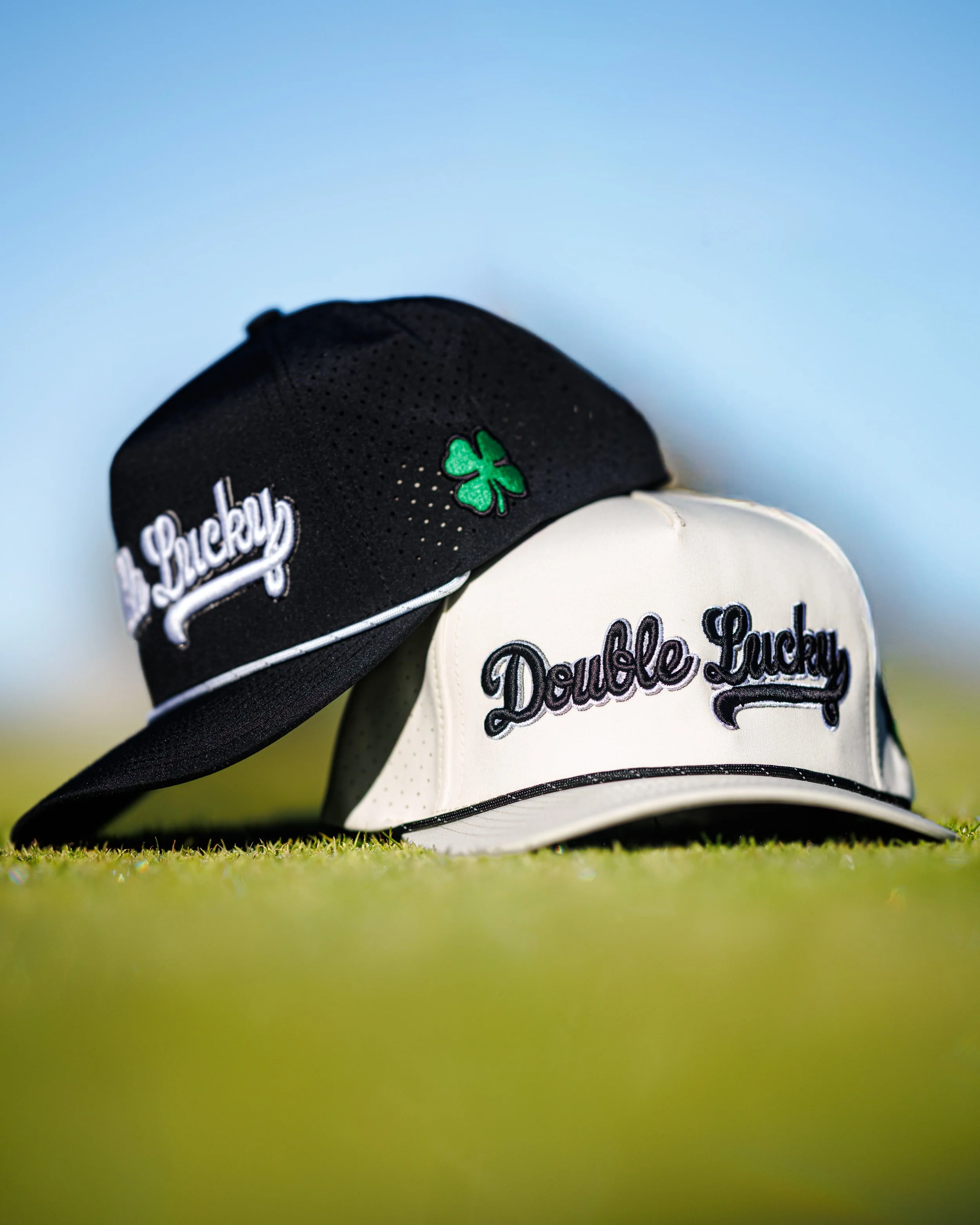 Two baseball caps, one black with a green shamrock and white embroidered text, and one white with black embroidered text, placed on grass outdoors under a bright blue sky.