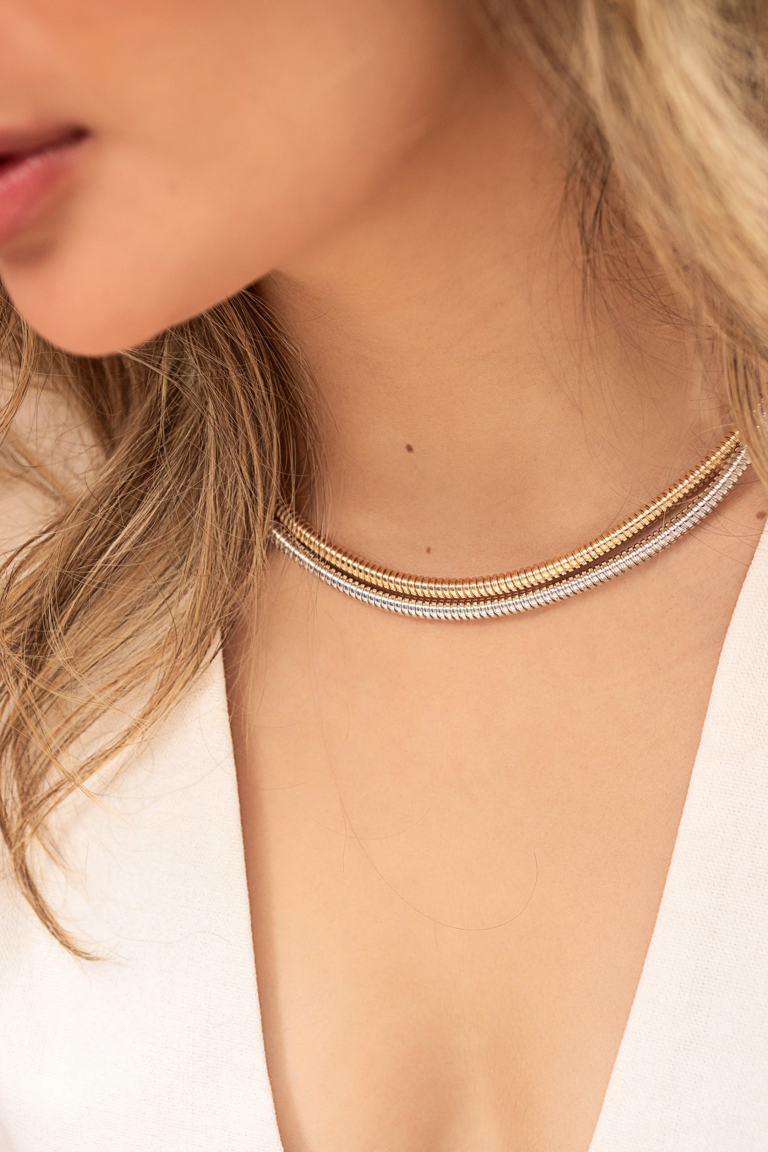 Cuello de mujer con collar de dos hilos finos, uno en oro y otro en plata, con cabello rubio y piel clara, vestida con prenda blanca.