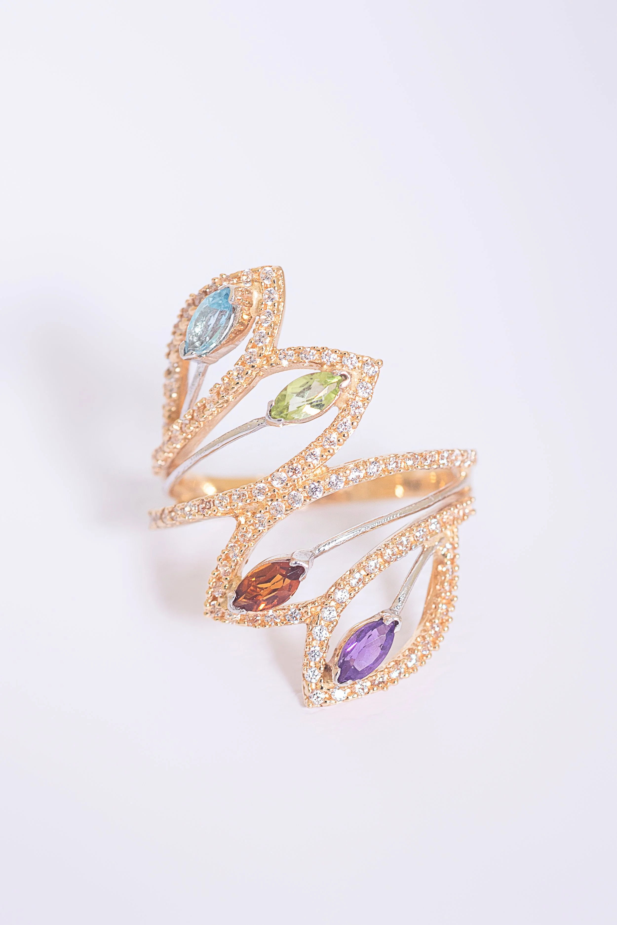 Anillo de joyería con diseño de hojas, decorado con múltiples piedras preciosas de diferentes colores y pequeños diamantes, en fondo blanco.