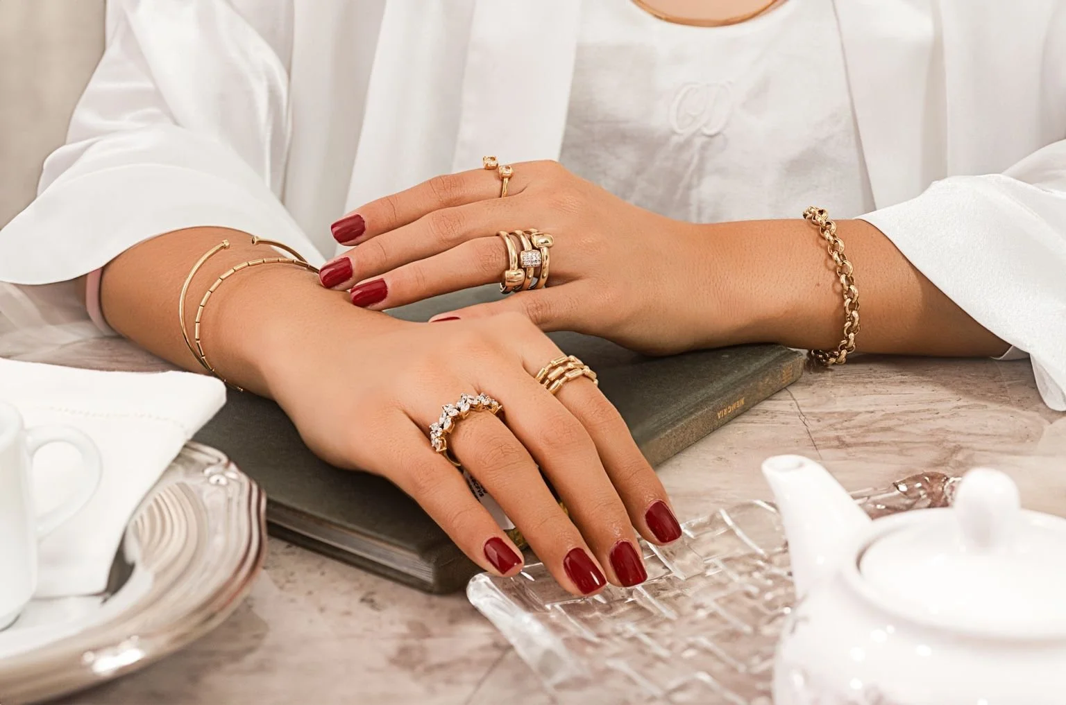 Manos con uñas pintadas de rojo, adornadas con varias joyas doradas y pendientes con piedras, descansando sobre un libro en una mesa de mármol, rodeadas de tazas y vajilla en un ambiente elegante.