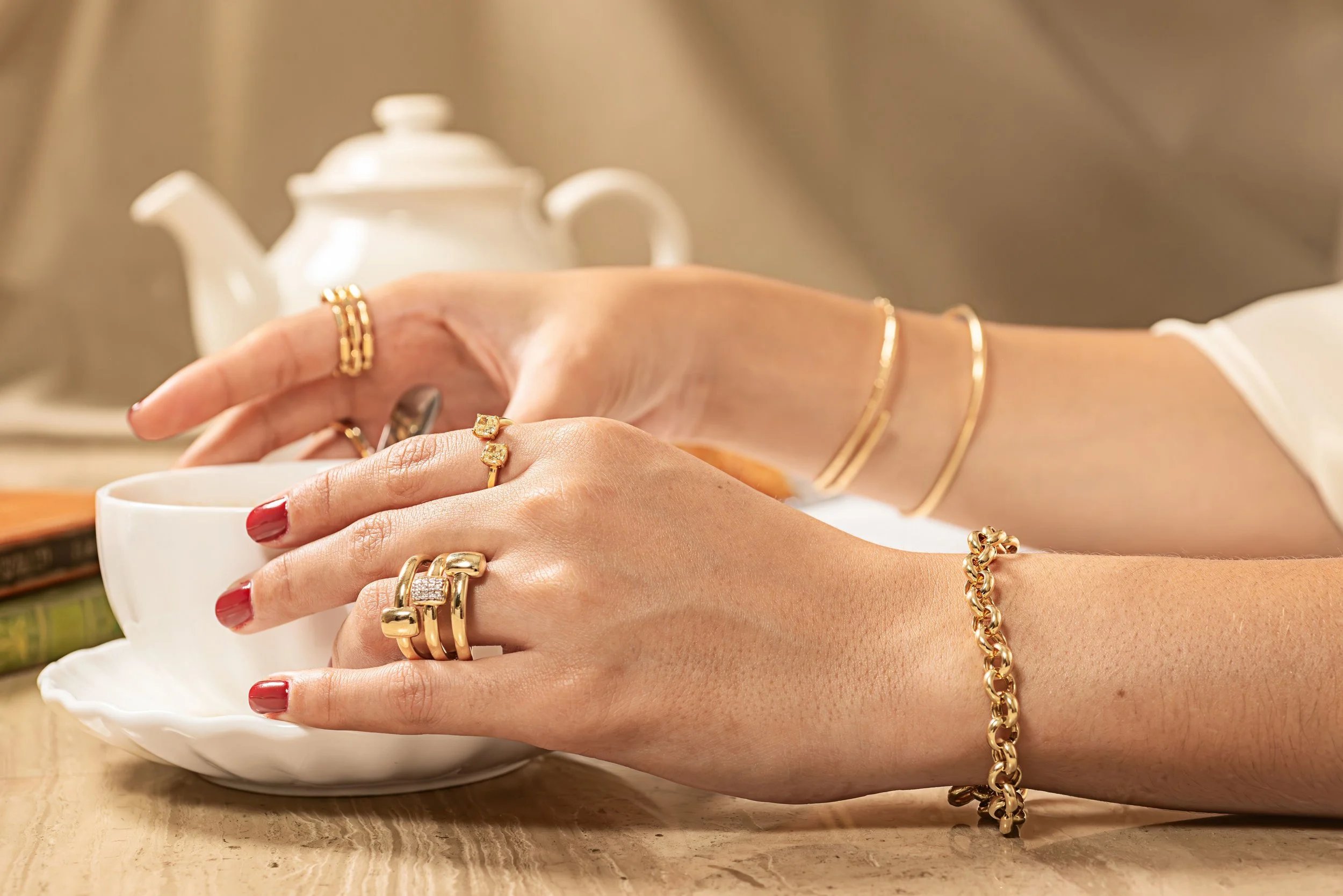 Manos con pulseras y anillos de oro, tocando una taza blanca en una mesa de madera, con una tetera blanca al fondo.